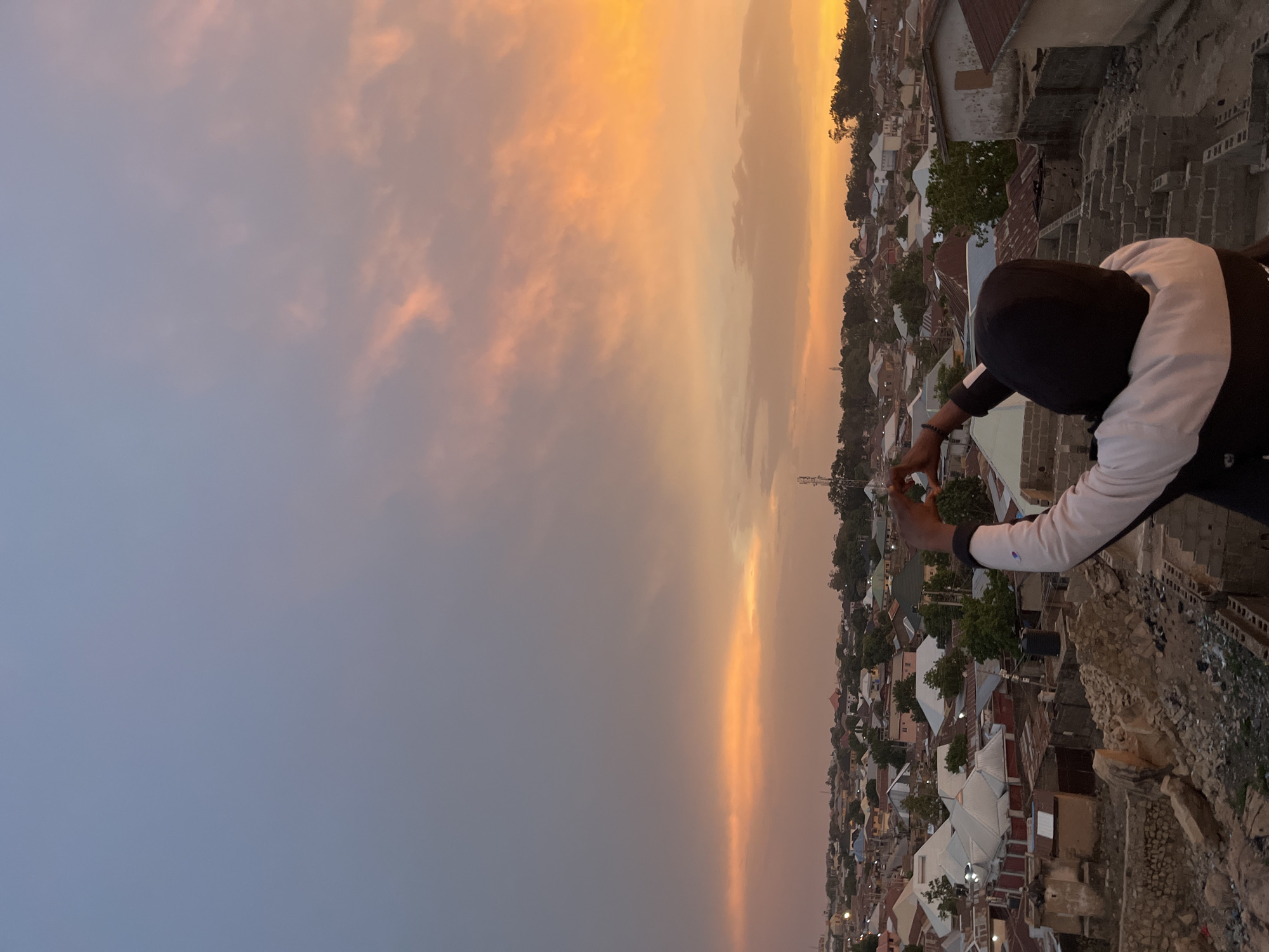 From an elevated viewpoint, the city of Jos, Nigeria, is captured during a sunset. The sky transitions from a blue-grey hue to a vibrant orange, with clouds tinged in shades of gold and pink. A person stands at the edge of the structure, clad in a black cap and a white shirt with black sleeves, their hands forming a heart shape towards the sunset. Below, the urban sprawl of Jos is visible, with rooftops, trees, and distant structures blending into the landscape. In the foreground, the edge of the building is rough and unfinished, made of light-colored stones and brick.