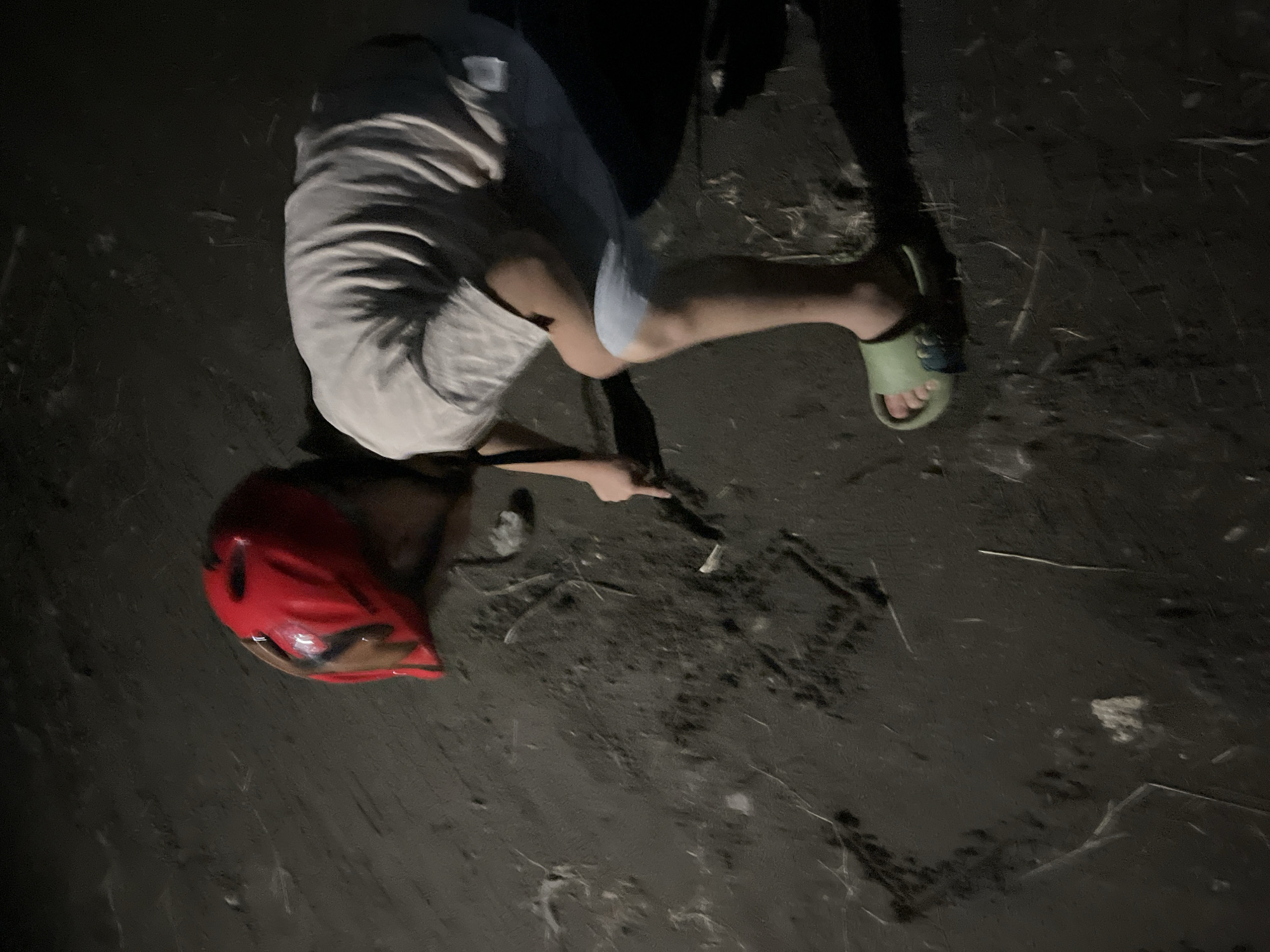 The image shows a child, wearing a red helmet and light-colored clothing, drawing a heart shape in the dark, sandy soil. The child is kneeling on the ground, with their right hand holding a stick or finger, and their left hand resting on the ground for balance. The child is wearing green sandals on their feet. The environment appears to be outdoors, possibly near a body of water, given the sandy texture of the ground. The lighting suggests it is either evening or night. There are no other individuals visible in the frame. The scene is devoid of any urban elements or landmarks that would specifically identify it as being in Poti, Georgia.