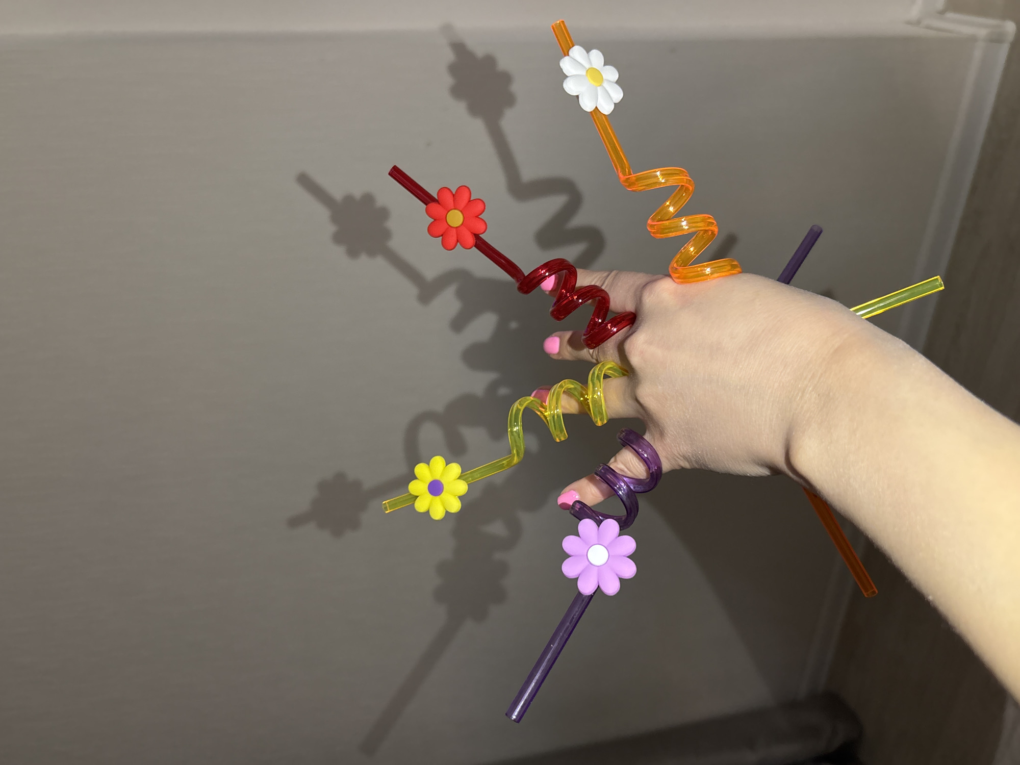 A hand holds five flower-adorned, curly straws of different colors (red, orange, purple, yellow, and orange again) with their shadows cast on a beige wall. The location is not discernible as Izhevsk, Russia.
