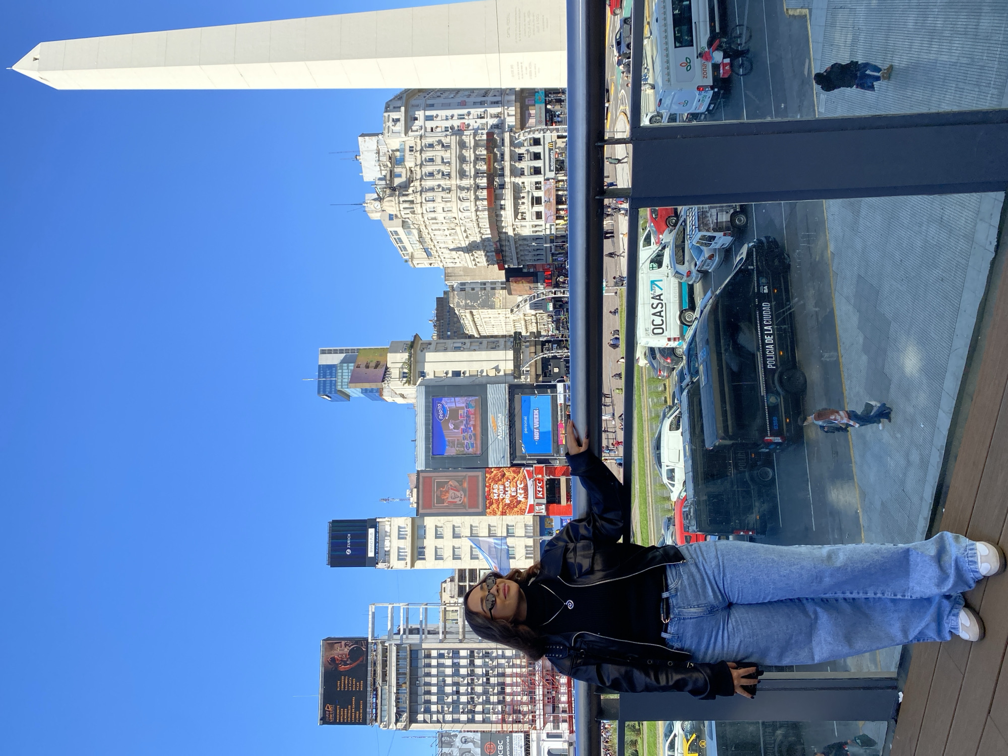 A medium shot depicts a young woman posing in front of a panoramic view of the city of Buenos Aires, Argentina, recognizable by the Obelisco monument towering in the distance against a clear, bright blue sky. The woman, wearing glasses, a black leather jacket, jeans, and white shoes, leans on a dark-colored railing. Below the railing, cars, buses, and vans—one bearing the name "OCASA" and another reading "POLICIA DE LA CIUDAD"—are visible on the street. Buildings line the cityscape, some featuring large advertisements and digital billboards. Pedestrians can be seen on the streets between the buildings and vehicles.
