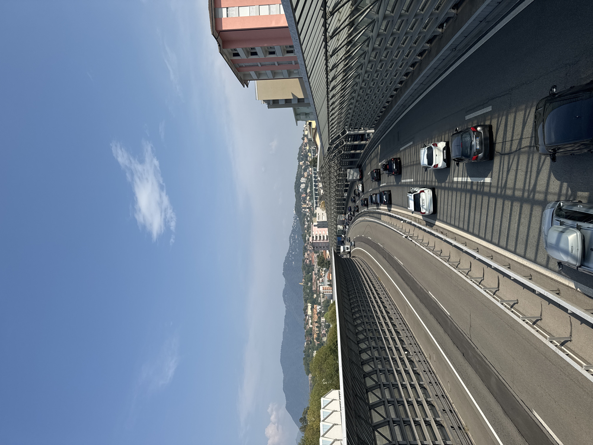 The image captures a section of the A2 motorway in Chiasso, Switzerland, during daytime. The perspective is from an elevated viewpoint, looking down onto the multi-lane highway. The road is congested with traffic, featuring numerous cars and a few trucks. On either side of the carriageway, there are extensive sound barrier structures, characterized by a metal framework supporting transparent panels and a solid wall base.
The urban landscape of Chiasso unfolds in the background. Buildings of varying heights and architectural styles are visible, including a prominent pink multi-story building with white window frames. Beyond the city, a range of hills and mountains forms the horizon, with one large mountain dominating the background. The sky is clear blue with a few wispy clouds. The lighting suggests it is a bright, sunny day, casting distinct shadows from the sound barriers onto the road and vehicles.