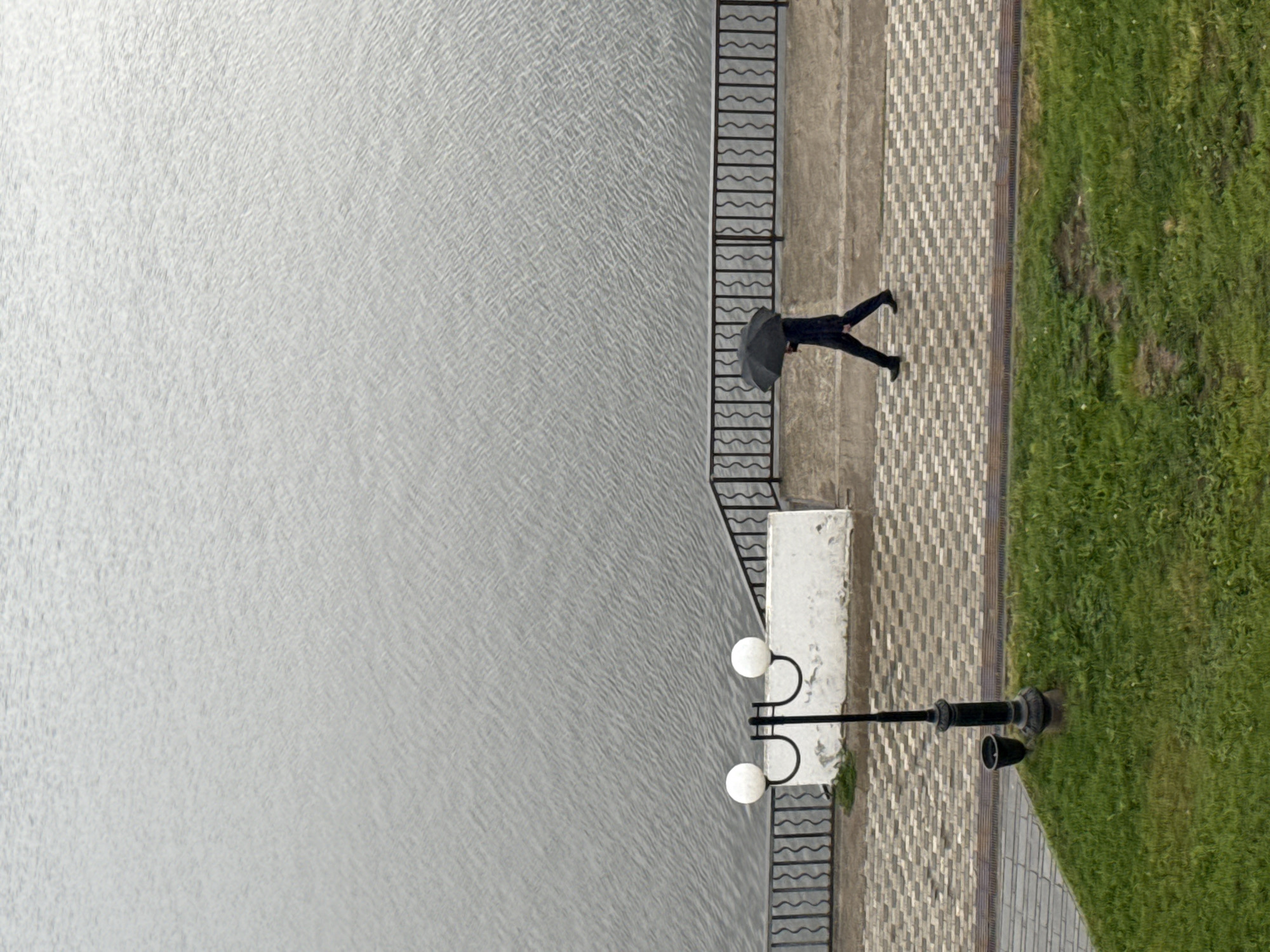 This high-angle, top-down shot captures a waterfront scene in Votkinsk, Russia, on an overcast day.
**Subjects:** The left side of the frame is dominated by a large body of water, likely a lake or river, displaying a finely rippled, textured surface in shades of grey. A dark, ornate metal railing with wavy horizontal bars separates the water from the land. To the right of the railing is a paved walking path, constructed with light-colored bricks arranged in a herringbone pattern. Further to the right, a strip of vibrant green grass borders the path.
A solitary person, dressed in dark clothing, is visible walking along the paved path, heading towards the upper part of the frame. They are holding an open black umbrella over their head. Along the path, there are two distinct streetlights. One features a dark post with two spherical white globes mounted on curved arms, positioned on a white rectangular concrete base near the railing. Another streetlight with a single globe is visible further along the path.
**Activity or event taking place:** The primary activity depicted is a person walking along a waterfront promenade. The open umbrella strongly suggests protection from rain, consistent with the diffused, even lighting that indicates an overcast sky.
**Notable details:** The image is characterized by its high-angle perspective, looking directly down onto the scene, which creates a flattened, almost geometric composition. The weather appears to be overcast or rainy, given the umbrella and the soft, shadowless lighting. The location details—the paved path, decorative railing, streetlights, and neatly maintained grass—suggest a public park or urban embankment along the water's edge in Votkinsk, Russia.
**Visible text:** No discernible text is visible in the media file.