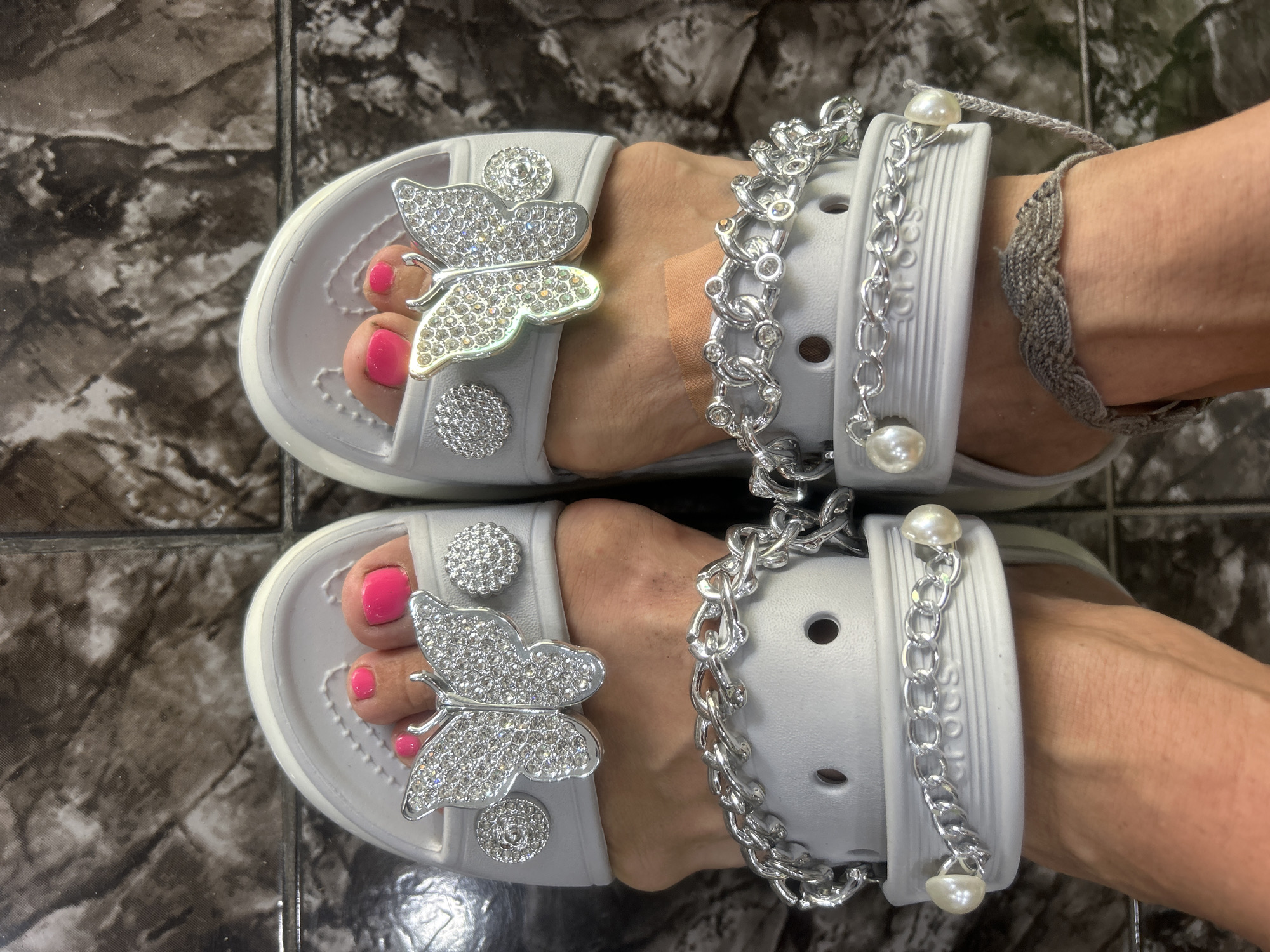The image shows two feet wearing grey Crocs sandals adorned with rhinestone butterfly and flower embellishments, as well as silver chains and pearl accents attached to the sandal straps. The toenails are painted bright pink. An anklet is worn on one ankle. The sandals rest on a dark, patterned tile floor. The location is inferred based on the user prompt as Nha Trang, Vietnam. The specific setting is not discernible from the image, but it appears to be indoors, possibly a home or commercial space.