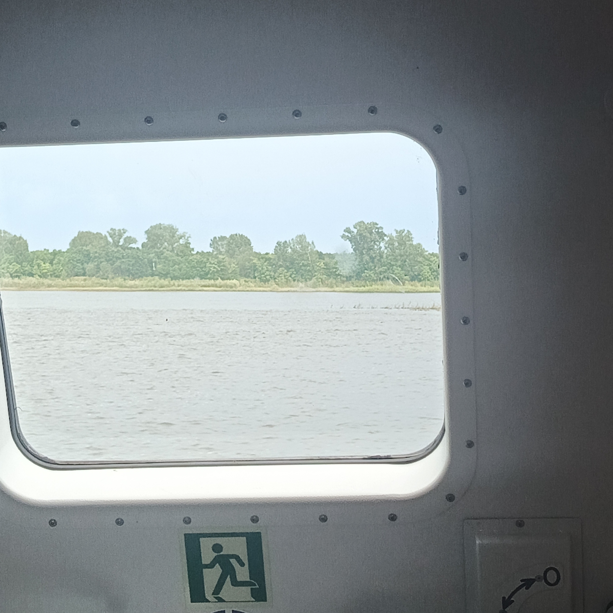 This image captures a view through a boat's window, looking out onto a body of water with a tree-lined shore in the distance. The window is an oval shape, framed by a white, textured interior. Several small, dark circular rivets are visible along the edge of the interior frame. Below the window, a green and white "exit" sign is mounted on the wall, featuring a silhouette of a running person. To the right of the exit sign, a small, recessed panel with circular markings and arrows suggests a control or indicator. The water is calm with a slightly choppy surface, reflecting the overcast sky. The distant shore is dense with various shades of green trees. The location of Nechkino, Russia, is not discernable from the visual information presented in the image.
