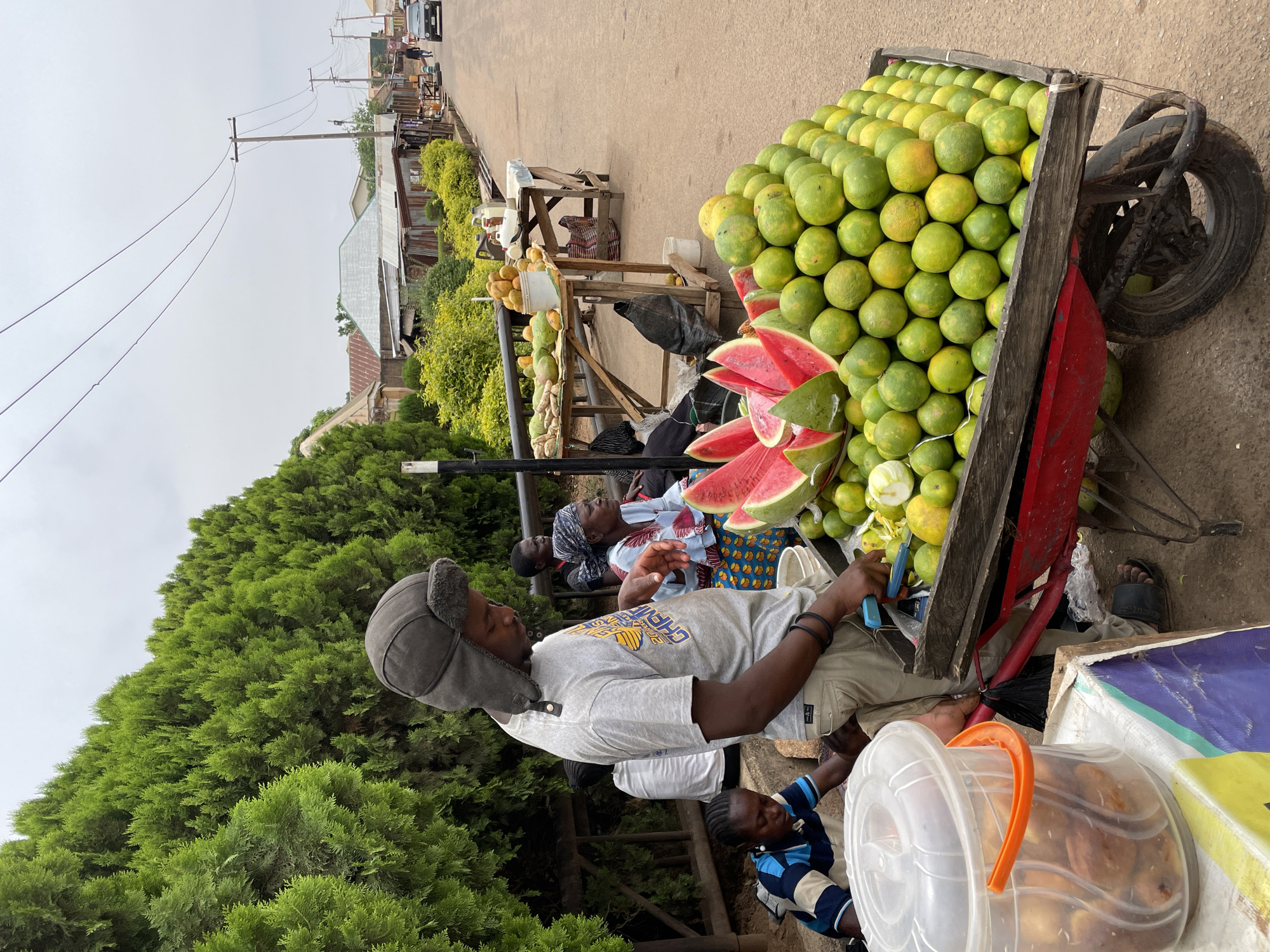 This image depicts a bustling roadside fruit market scene in Jos, Nigeria, under an overcast sky. In the foreground, a man wearing a grey t-shirt with a "GHANA" logo and a grey ear-flap hat stands beside a large wooden pushcart. The cart, which has a red frame and a visible wheel, is overflowing with a substantial quantity of green and yellow citrus fruits, likely oranges or grapefruits, and several bright red slices of watermelon. The man appears to be attending to the stall, holding a blue object in his hand, possibly a phone or a cutting tool.
Behind him, several other individuals are present, suggesting both vendors and customers. A woman in a patterned dress and headscarf, along with others, are seated or standing near additional wooden stalls that display other fruits such as mangoes and potentially papayas. In the immediate foreground, a clear plastic bucket with an orange handle holds what appear to be snacks or baked goods.
The market is situated along a road, which is bordered by lush green bushes and trees. Utility poles with overhead lines are prominent against the cloudy sky, and in the distance, buildings with corrugated metal roofs can be seen. The scene captures the daily activity of a vibrant local market, with people engaged in buying and selling fresh produce.