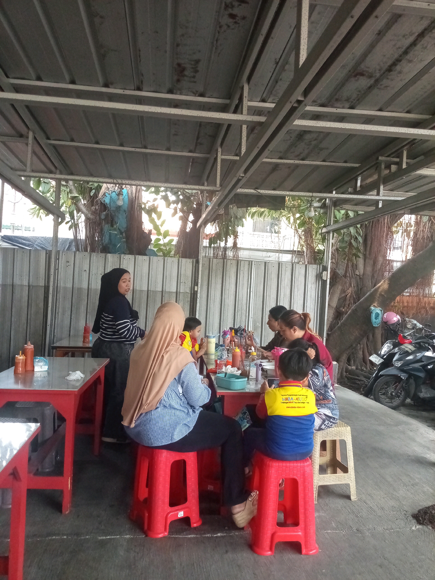 The image depicts an outdoor dining area at a roadside eatery in Bekasi, Indonesia. A group of people, including women and children, are seated at tables, seemingly enjoying a meal. The setting is characterized by a metal roof structure supported by beams, providing shade. Red plastic tables and stools are arranged on a concrete floor. Various food condiments, such as ketchup bottles, are visible on the tables. In the background, a large tree with exposed roots and a corrugated metal fence are present. Several motorcycles are parked to the right of the scene. The overall atmosphere suggests a casual and communal dining experience.