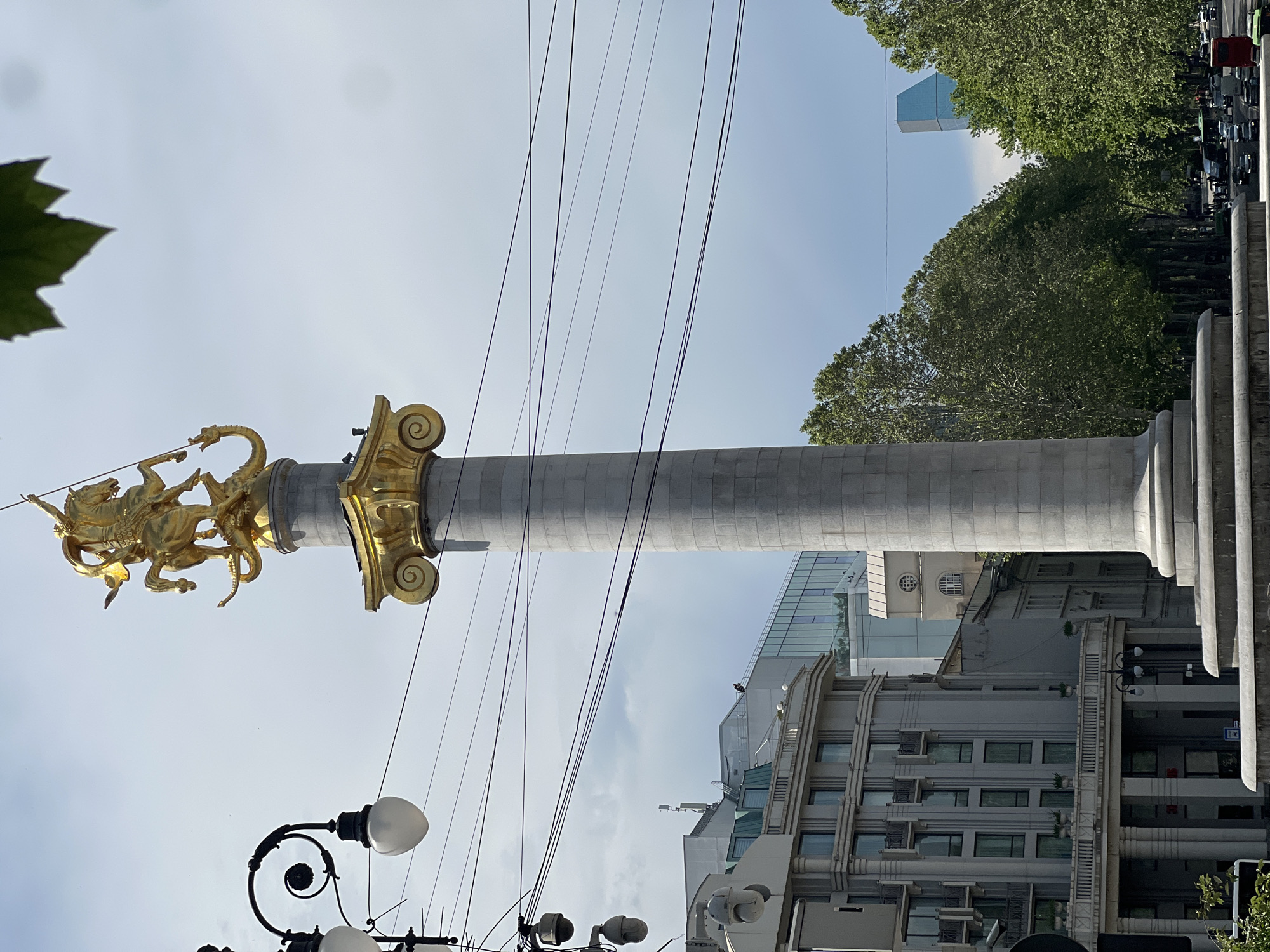 The image shows the St. George statue atop the column in Freedom Square in Tbilisi, Georgia. The statue depicts St. George slaying the dragon. The column is made of stone. Utility wires are visible in the foreground. Buildings and trees are visible in the background. The sky is partially cloudy. A black lamppost with white glass globes is in the foreground.