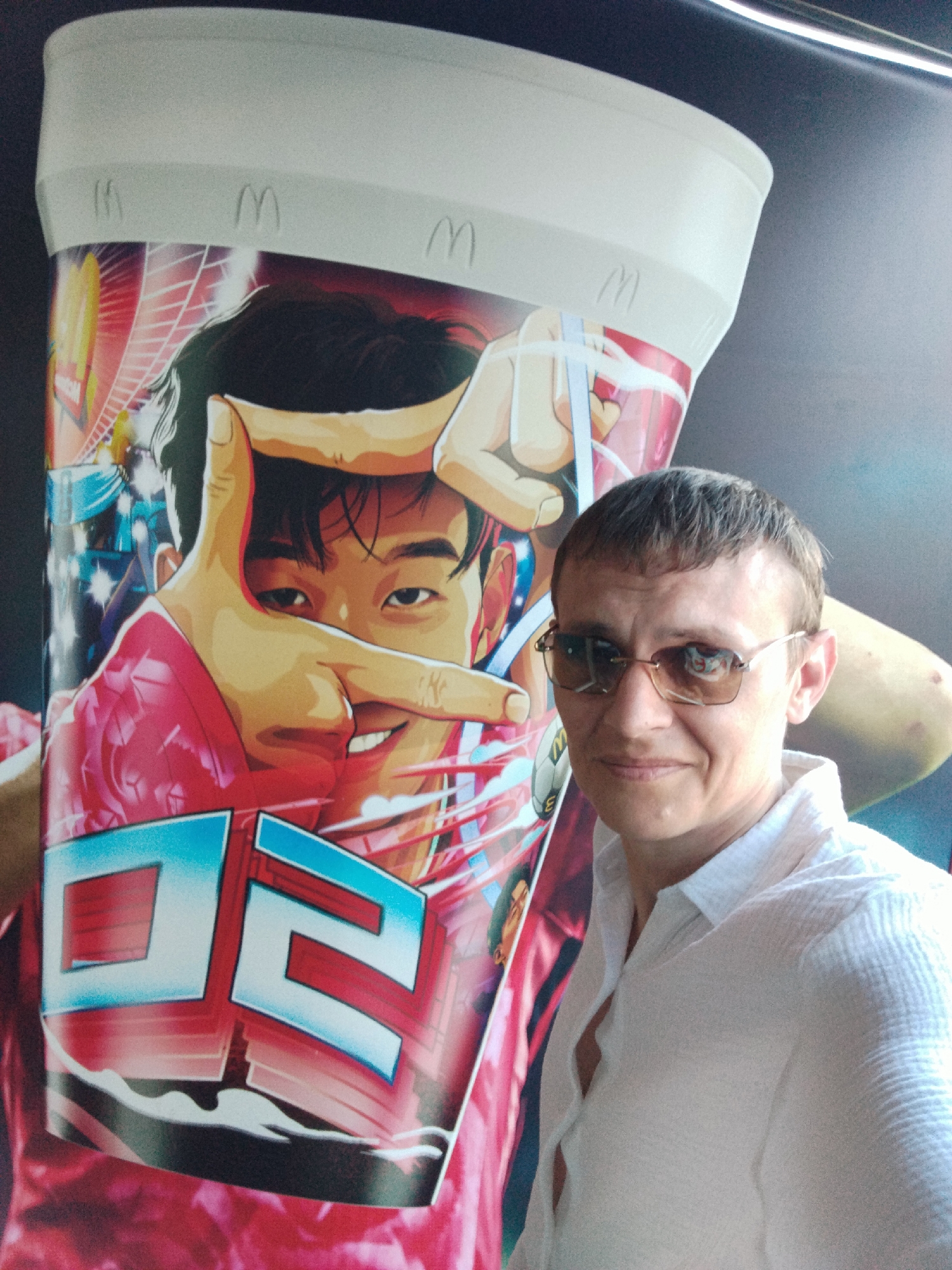The image shows a man in a white collared shirt and sunglasses standing in front of a large McDonald's cup advertisement featuring an anime-style illustration of a person. The advertisement is a bright and colorful graphic with the McDonald's logo visible at the top. The man is smiling and looking at the camera. The background appears to be an indoor setting, possibly a McDonald's restaurant in Be'er Sheva, Israel.