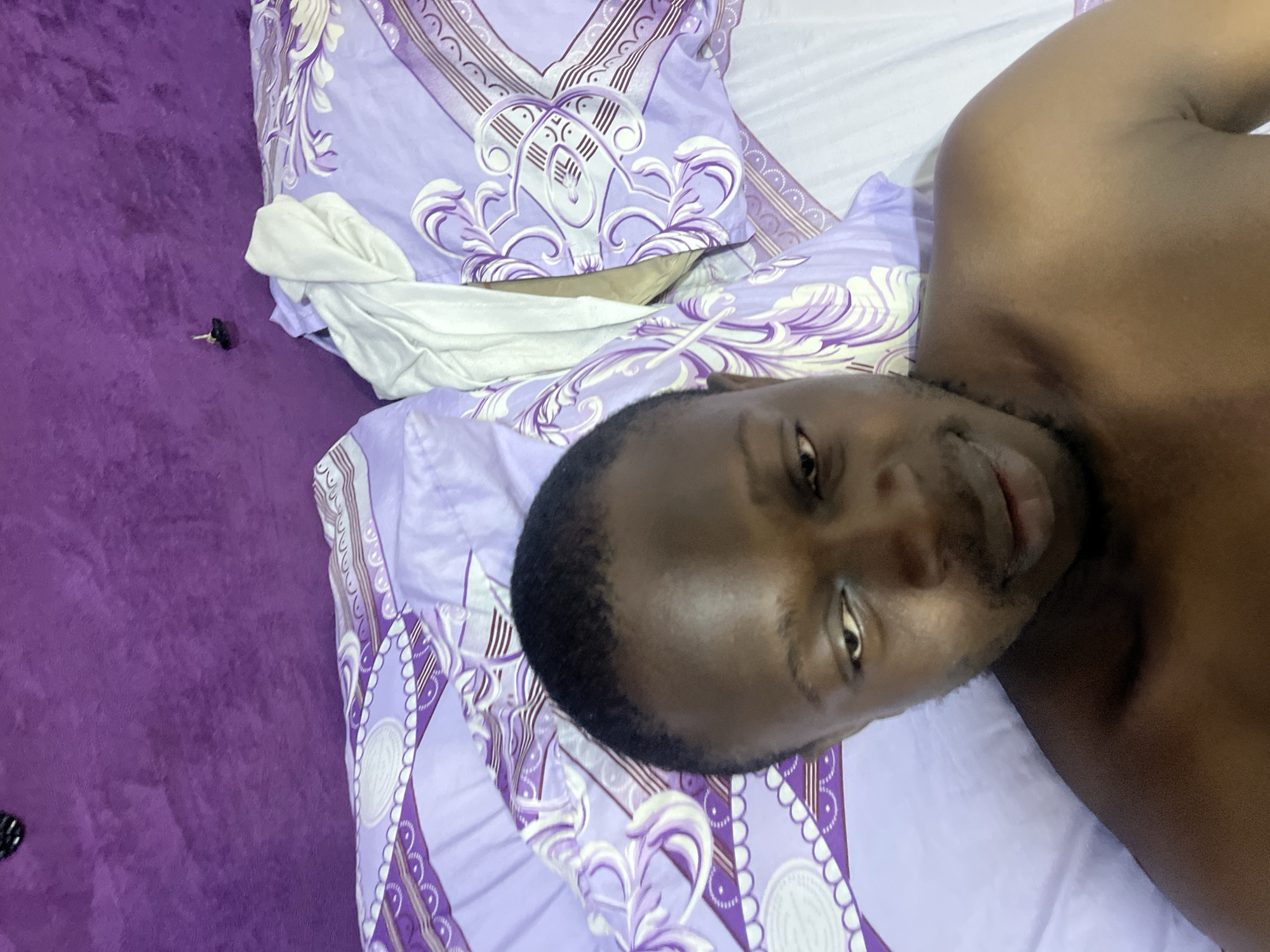 This image depicts a male individual with dark skin, short dark hair, and light facial hair, shirtless, lying in a bed within an indoor setting in Abuja, Nigeria. He is positioned from the chest up, looking downwards and slightly to his right. Behind him is a purple, plush, or upholstered headboard or wall. Two pillows with light purple and white patterned pillowcases, featuring abstract swirling designs, circular elements, and striped borders, are arranged on the bed. A crumpled white piece of fabric is placed between the pillows. A small, dark, irregular object rests on the purple surface near the top center of the frame. The image is taken from a slightly elevated, close-up perspective.