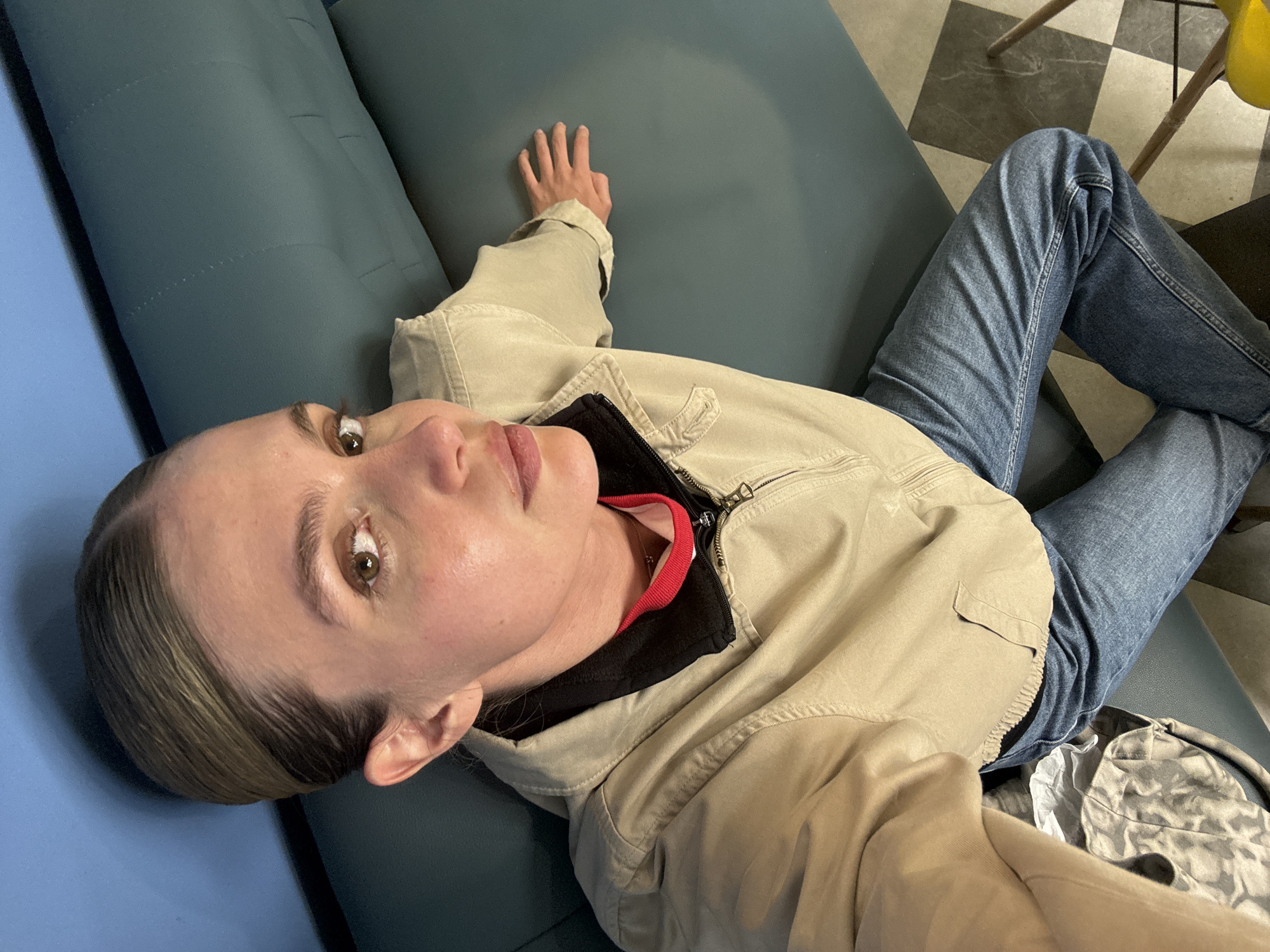 A woman is lying on a blue couch indoors. She has fair skin, light brown hair pulled back, and is wearing a beige jacket over a black top with a red collar, and blue jeans. Her right hand is extended on the couch. A patterned floor is visible in the background. This photograph was taken in Tbilisi, Georgia.
