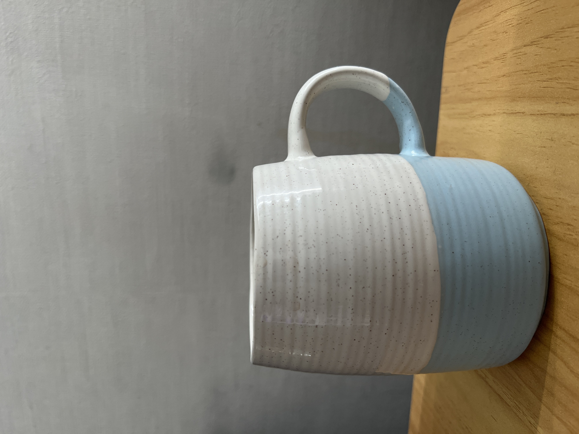 The image shows a two-toned mug with a handle. The mug is primarily white with dark speckles on the left and a lighter blue on the right, with the handle also partly blue. The mug appears to be resting on a light brown, possibly wooden surface. The background behind the mug is a plain, light gray wall. The scene lacks any discernible features that would identify it as being located in Saint Petersburg, Russia.