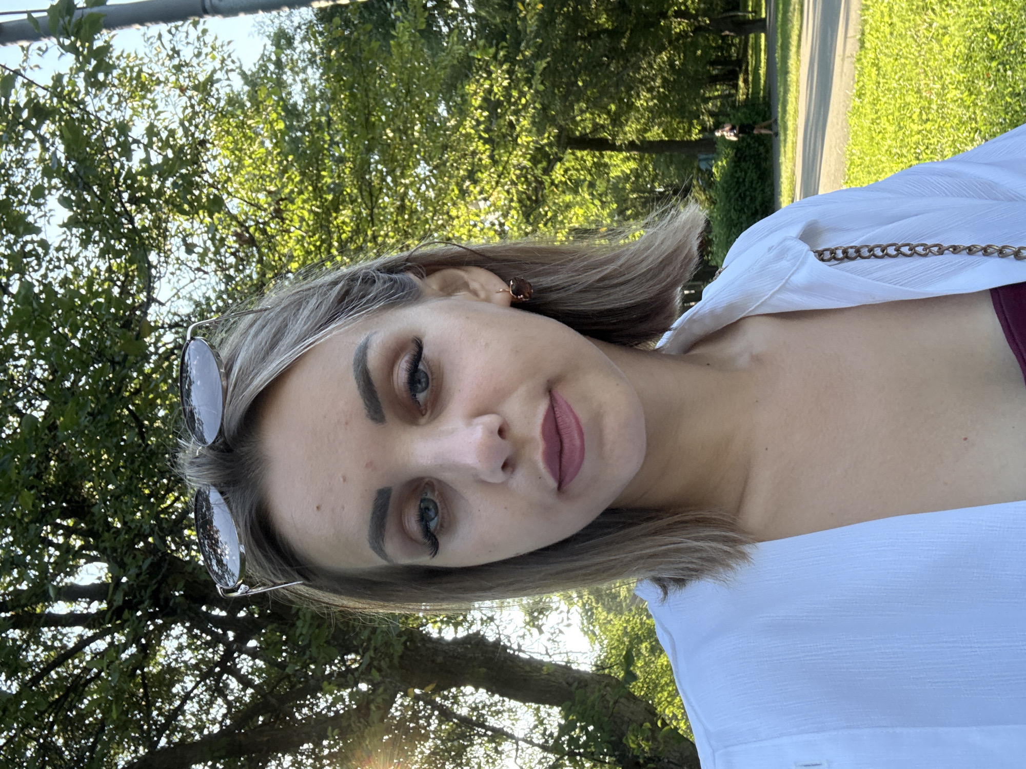 In this portrait-style image taken outdoors, a young woman with blonde hair is the primary subject. She is positioned in the foreground, facing the camera, and her gaze is directed slightly to the left. She is wearing a white button-up shirt, partially unbuttoned, revealing a glimpse of a burgundy garment underneath. A gold chain accessory is visible on her shoulder, attached to her clothing. Her makeup includes noticeable eyeliner and mascara, and her lips are painted with a matte lipstick. She is also wearing large, reflective sunglasses pushed up onto her head.
The background suggests an urban park setting, likely in Yuzhno-Sakhalinsk, Russia, indicated by the presence of lush green trees with dappled sunlight filtering through, and a paved pathway in the middle ground. A person can be seen walking in the distance on this path. The overall lighting is bright, with sunlight creating strong highlights and shadows, suggesting it's daytime. The presence of the trees, pathway, and distant figure are consistent with a park environment.