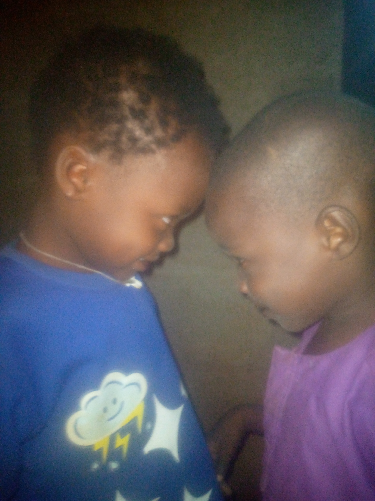 This close-up image captures two young children, likely toddlers or preschoolers, with dark skin tones, sharing a tender moment. They are positioned facing each other, their foreheads gently touching as they both look downwards, suggesting affection, shyness, or focused interaction.
The child on the left is wearing a vibrant blue long-sleeved shirt, which features a prominent white graphic of a smiling cloud with yellow lightning bolts underneath it. A simple, thin white necklace is visible around this child's neck. Their hair is short and textured, possibly styled with small braids or patterns visible on the scalp.
The child on the right is dressed in a plain purple shirt and has very short, possibly shaved hair. Both children's faces are partially obscured by their pose and downward gaze, but the overall impression is one of quiet connection.
The scene appears to be set indoors against a dark, indistinct background, which could be a wall. The lighting is dim and soft, contributing to the intimate atmosphere but also resulting in a slightly blurry and grainy image quality. No specific location cues are visible beyond an indoor setting. There is no visible text in the image.