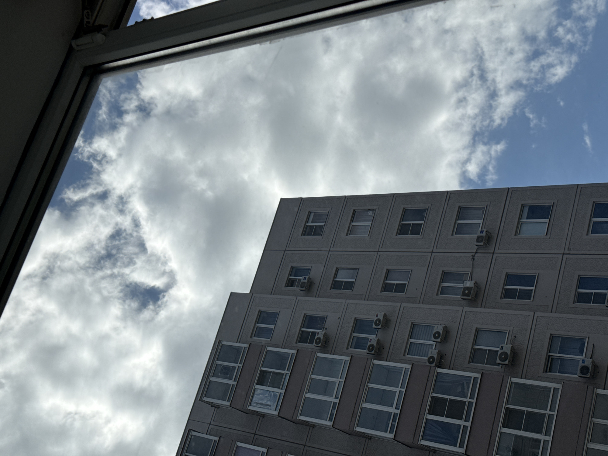 A low-angle shot taken from inside a building through an open window shows a portion of a multi-story, concrete building facade against a cloudy sky. The building, possibly located in an urban area of Rostov-on-Don, Russia, has rows of rectangular windows, each fitted with air conditioning units beneath. The window frame through which the photo is taken is visible in the lower left, and the rest of the view is filled with the building and the sky. The sky is overcast with varying shades of grey and white clouds interspersed with patches of blue.
