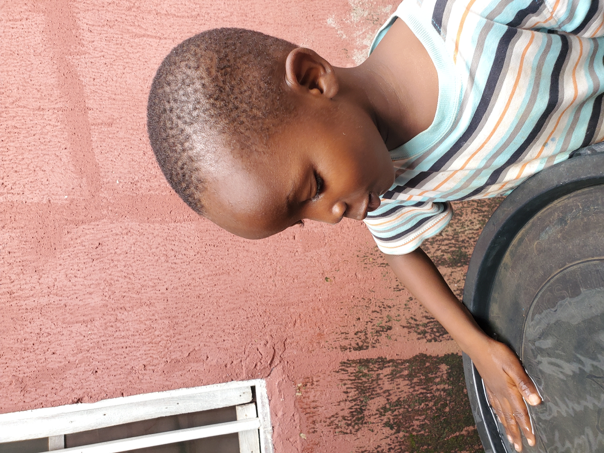 The image features a young boy, viewed from a high angle, with his head tilted downwards and to the right. He is wearing a striped t-shirt with horizontal bands of blue, white, orange, and black. His right arm extends towards a large, dark, round basin filled with water, and his hand is submerged in the water. The boy's skin tone is dark.
The background consists of a textured, reddish-pink wall with some green mossy patches and dark shadows on the ground. In the lower left corner, a portion of a white wooden structure, possibly part of a window or a vent, is visible. The lighting suggests it is daytime, possibly outdoors, given the shadows. The boy's expression is difficult to ascertain due to the angle and his focus on the water. There is no visible text in the image.
