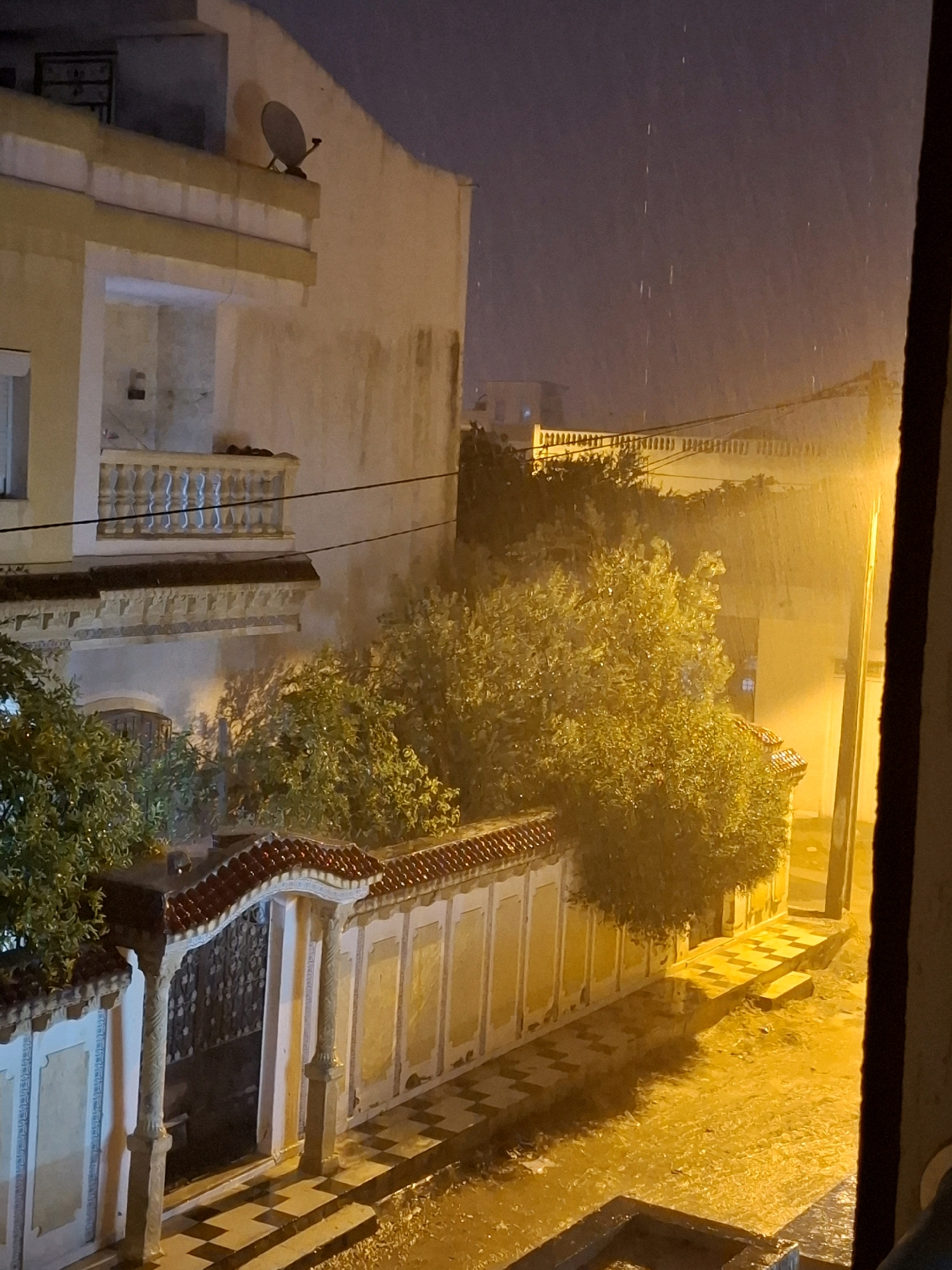 This is a nighttime, slightly blurry, photograph taken from an elevated perspective, looking out a window onto a street scene in Bennane, Tunisia. The scene depicts a building with a light-colored facade, a balcony with white ornate railings, and a satellite dish mounted on the roof. In front of the building is a courtyard or street area with a patterned tiled walkway and a low wall with decorative elements. A dense, green bush is visible, illuminated by a bright yellow light source, likely a streetlamp, which also casts a strong yellow glow on the wet pavement and the surrounding area. The rain is evident, with visible streaks of water falling from the dark sky, and puddles forming on the ground, reflecting the artificial light. The overall atmosphere is that of a stormy, wet night in an urban or semi-urban environment.