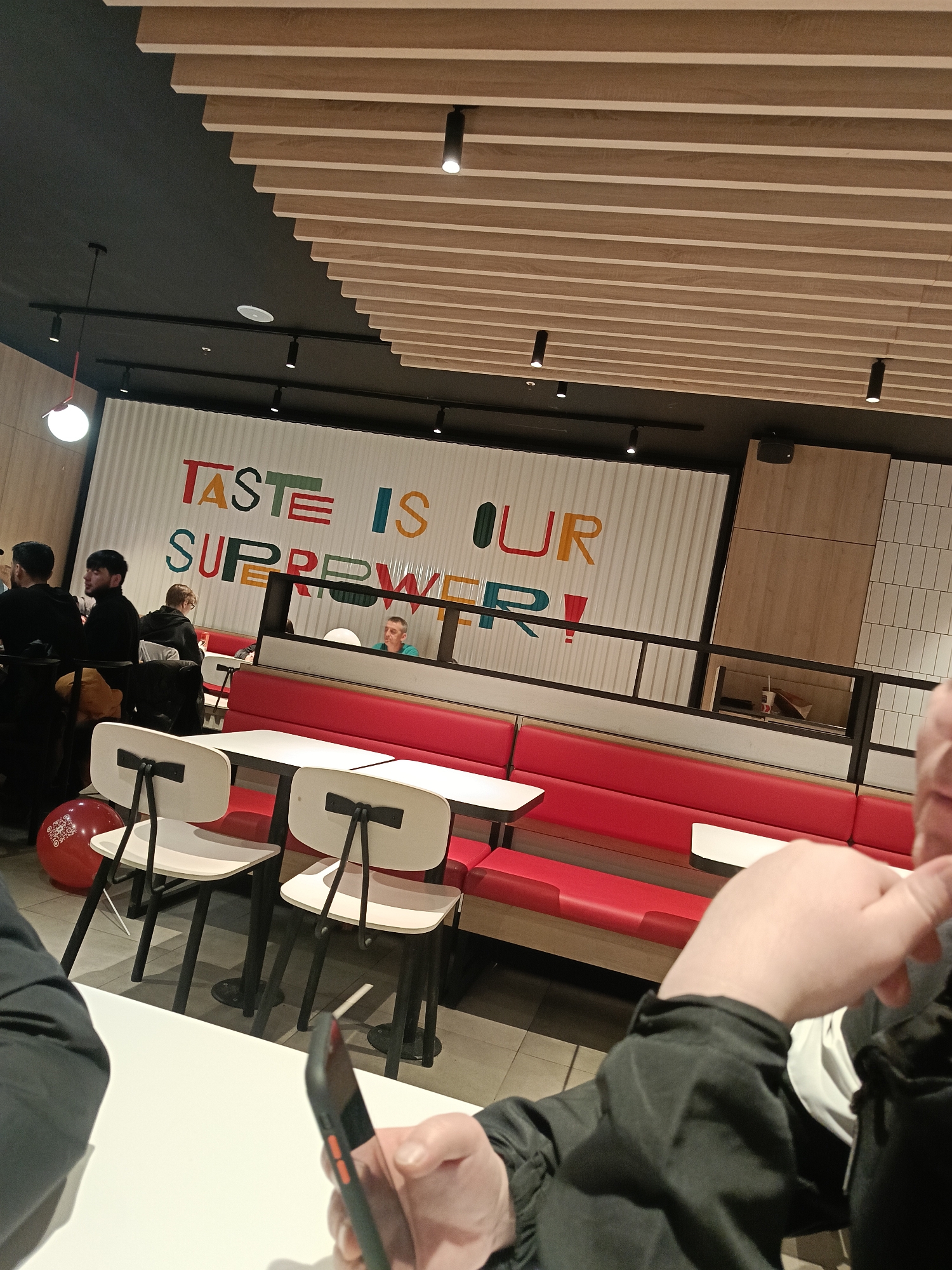 The image shows the interior of a McDonald's restaurant in Izhevsk, Russia. A large sign reading "TASTE IS OUR SUPERPOWER!" is visible on a white corrugated wall behind several occupied tables. Some people are sitting at tables, while others are waiting in line. There are red booths with white tables and black chairs with white backs. The ceiling is black with wooden slats. A red balloon is on the floor beside one of the chairs. A person in the foreground is holding a black smartphone. The interior is well-lit with a combination of overhead lighting fixtures.