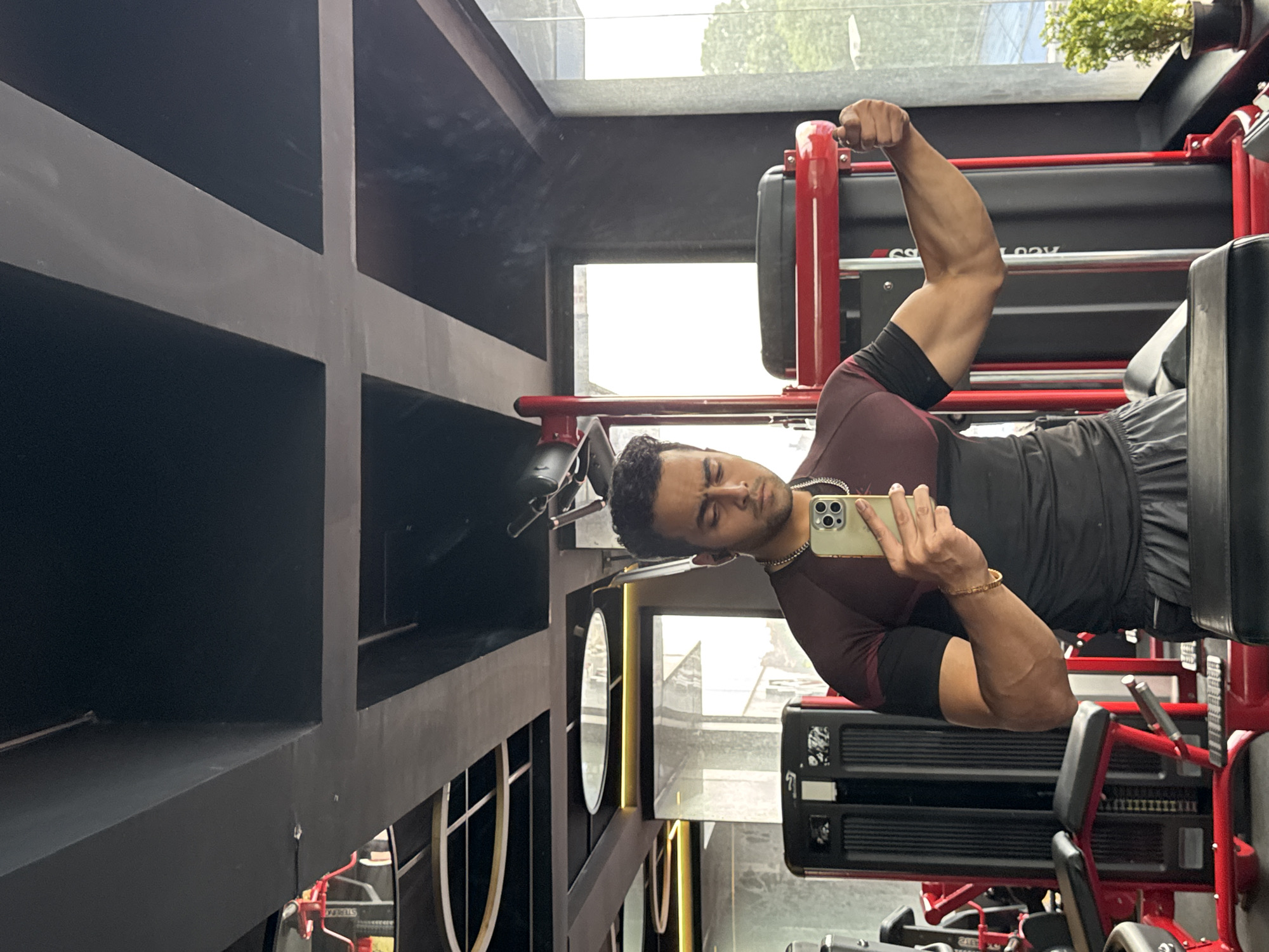 A man takes a mirror selfie in a gym. He is wearing a maroon and black t-shirt and grey shorts. He is flexing his bicep. The gym appears to have modern equipment and a large window with a view of trees and buildings. The ceiling has a unique design with black and gray panels and recessed lighting. The gym is likely located in a city, possibly Kolkata, India, given the context provided.