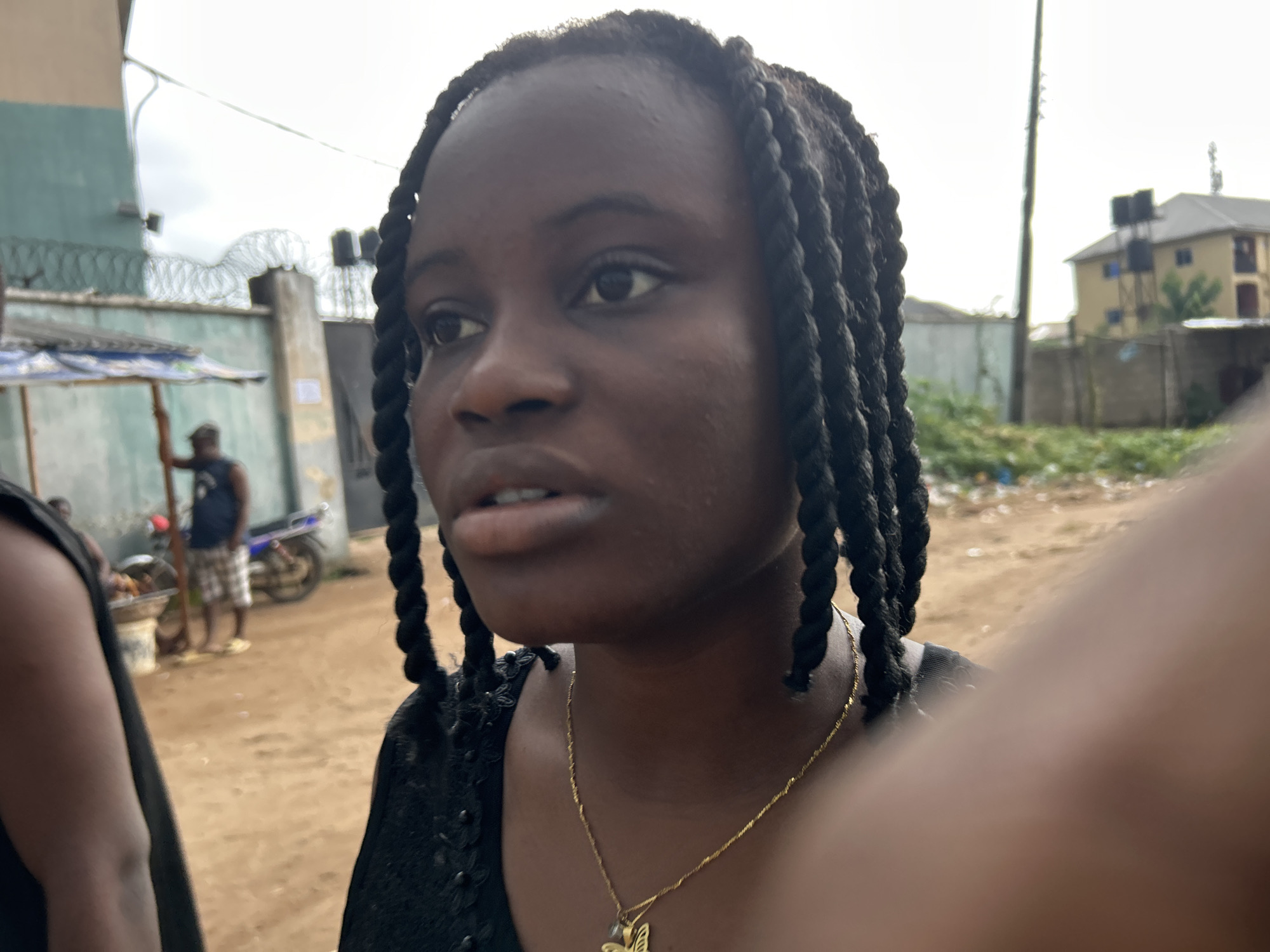 A close-up, selfie-style shot focuses on a young Black woman with braided hair, wearing a black top and a gold chain with a butterfly pendant. Her face is turned to the left, her expression unreadable. The background suggests an outdoor, informal setting in Ihiagwa, Nigeria, with dirt ground and a pale, overcast sky.
Behind the woman, a muted green building is visible on the left, partially obscured by a makeshift stall with a blue and white striped awning. A man in a dark t-shirt and plaid shorts stands near the stall, facing away from the camera, with a motorcycle parked behind him. Further back, a grey concrete wall topped with barbed wire runs horizontally across the frame. In the distance, a yellow building with a dark roof and water tanks on a raised structure can be seen, along with a tall pole that might be a utility pole. The ground is covered in dirt and scattered debris. The overall impression is one of a casual, everyday scene in a Nigerian community.