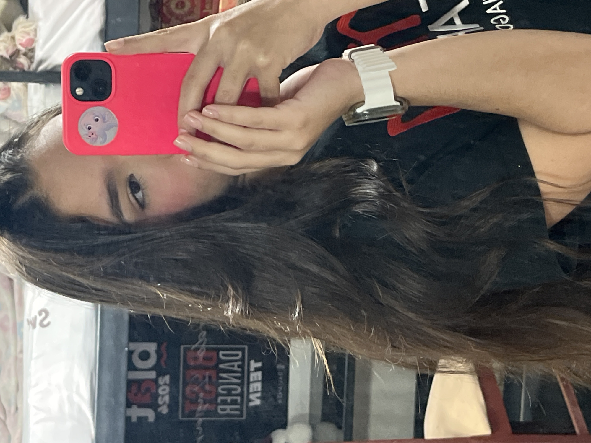 A young woman with long, dark, wavy hair is taking a selfie in Juan Griego, Venezuela. She is wearing a black t-shirt and a white smartwatch on her left wrist. She is holding a bright pink smartphone with a circular sticker of two cartoon octopuses on the back. In the background, reflected in a mirror, are signs with text that appears to be in English, including "ASOS" and "BEST DANCER TEEN". The environment appears to be indoors, possibly a bedroom or a common area, with a bed visible in the upper portion of the image.
