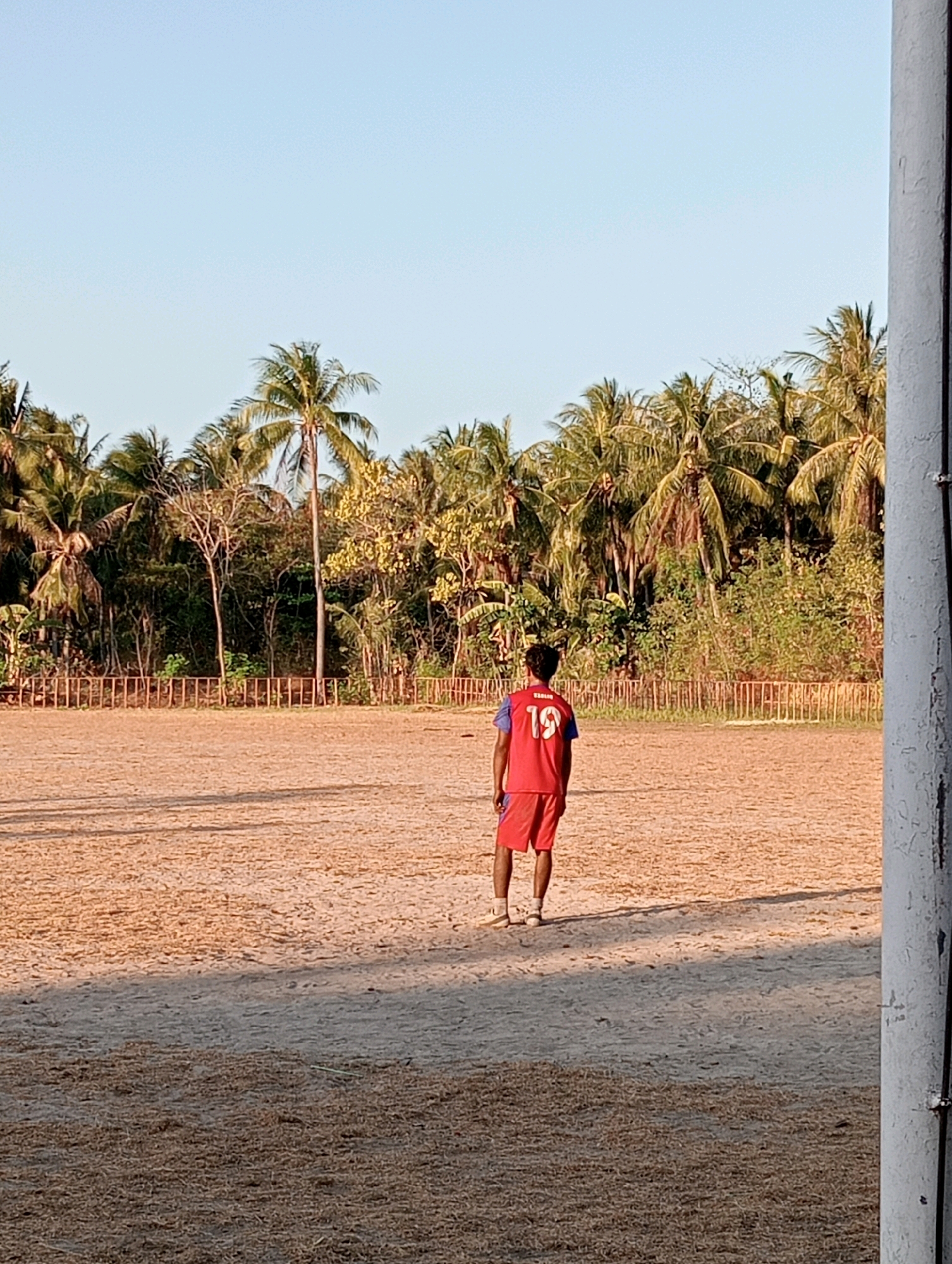 The image depicts a young person wearing a red and blue athletic jersey with the number "19" on the back, and red shorts, standing on a sandy field. The person is facing away from the camera, looking towards a line of palm trees and dense vegetation that forms the background. A thin wooden fence runs horizontally across the middle ground of the image, separating the sandy field from the vegetated area. The sky above is clear and light blue. The setting appears to be an outdoor sports field, possibly in a tropical or subtropical region. The presence of palm trees suggests a location like Pagerungan Kecil, Indonesia. The sandy ground is uneven and shows shadows from an unseen light source, possibly the sun. To the right of the frame, a portion of a white, textured vertical pole is visible.