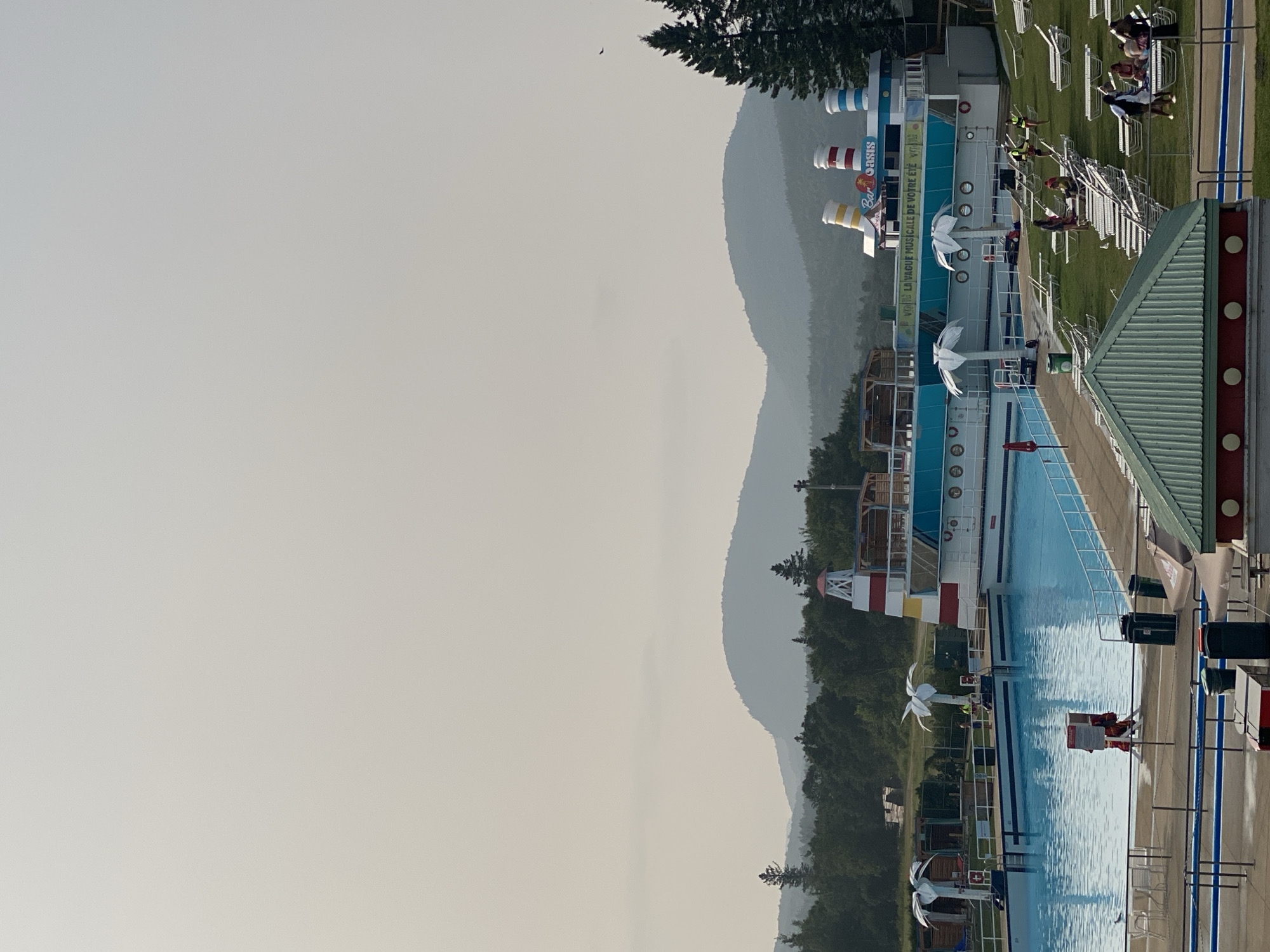 The image depicts an outdoor swimming pool complex, likely a water park, situated in Saint-Gabriel-de-Valcartier, Canada, given the prominent "Bar Oasis" sign which is a reference to the Village Vacances Valcartier. The scene is viewed from an elevated perspective, looking across the water.
In the foreground, a large, bright blue swimming pool dominates the left side of the frame. Several lounge chairs are arranged on the green grass surrounding the pool. In the middle ground, a large, white structure resembling a ship with blue accents and funnels is positioned adjacent to the pool. This structure features a lighthouse-like tower at one end and appears to house a bar, indicated by the "Bar Oasis" signage. Palm tree-like decorations are visible around the structure.
Several people are scattered throughout the scene, some lounging on chairs on the grass, while others appear to be near or in the pool. One individual in a red uniform, possibly a lifeguard, is standing by the edge of the pool. In the background, lush green trees and rolling, mist-covered mountains form a natural backdrop under a hazy, overcast sky. The overall impression is a leisure environment on a slightly obscured, possibly humid day.