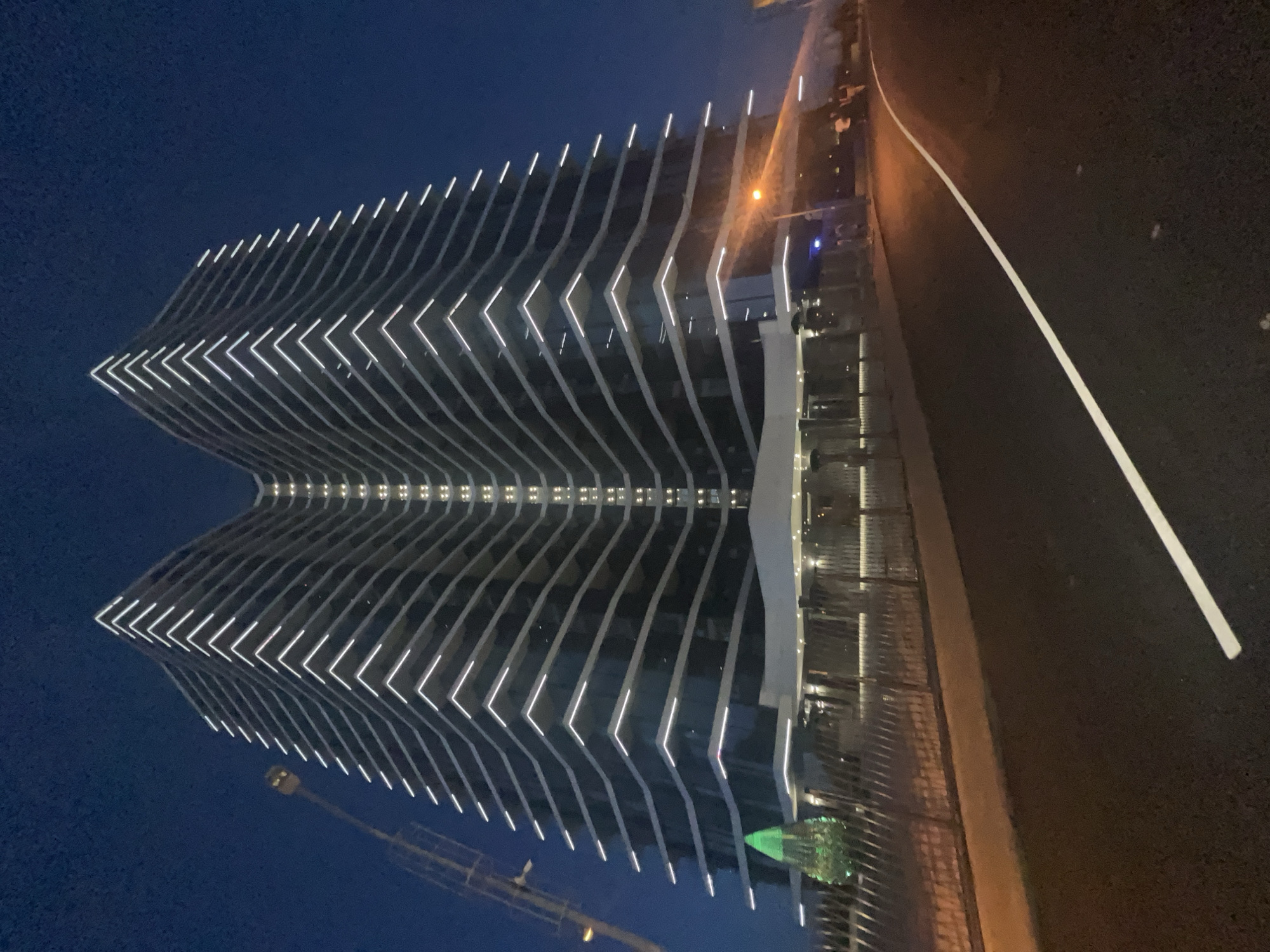 This image depicts a modern, tall building with a distinctive V-shaped facade illuminated by white LED strip lights along its horizontal edges. The building appears to be under construction or recently completed, given the presence of a crane in the background, visible on the left side of the frame. The lighting suggests it is evening or nighttime, with a deep blue sky. In the foreground, a dark asphalt road with a white lane marker curves to the right. A fence runs along the side of the building, and behind it, a few figures are visible. A solitary street lamp with an orange glow is present. There's a green, illuminated decorative tree to the left of the building's entrance. The overall setting appears to be an urban environment in Odesa, Ukraine, specifically on the site of the Odesa Business Port project, a mixed-use development that includes residential buildings and offices.