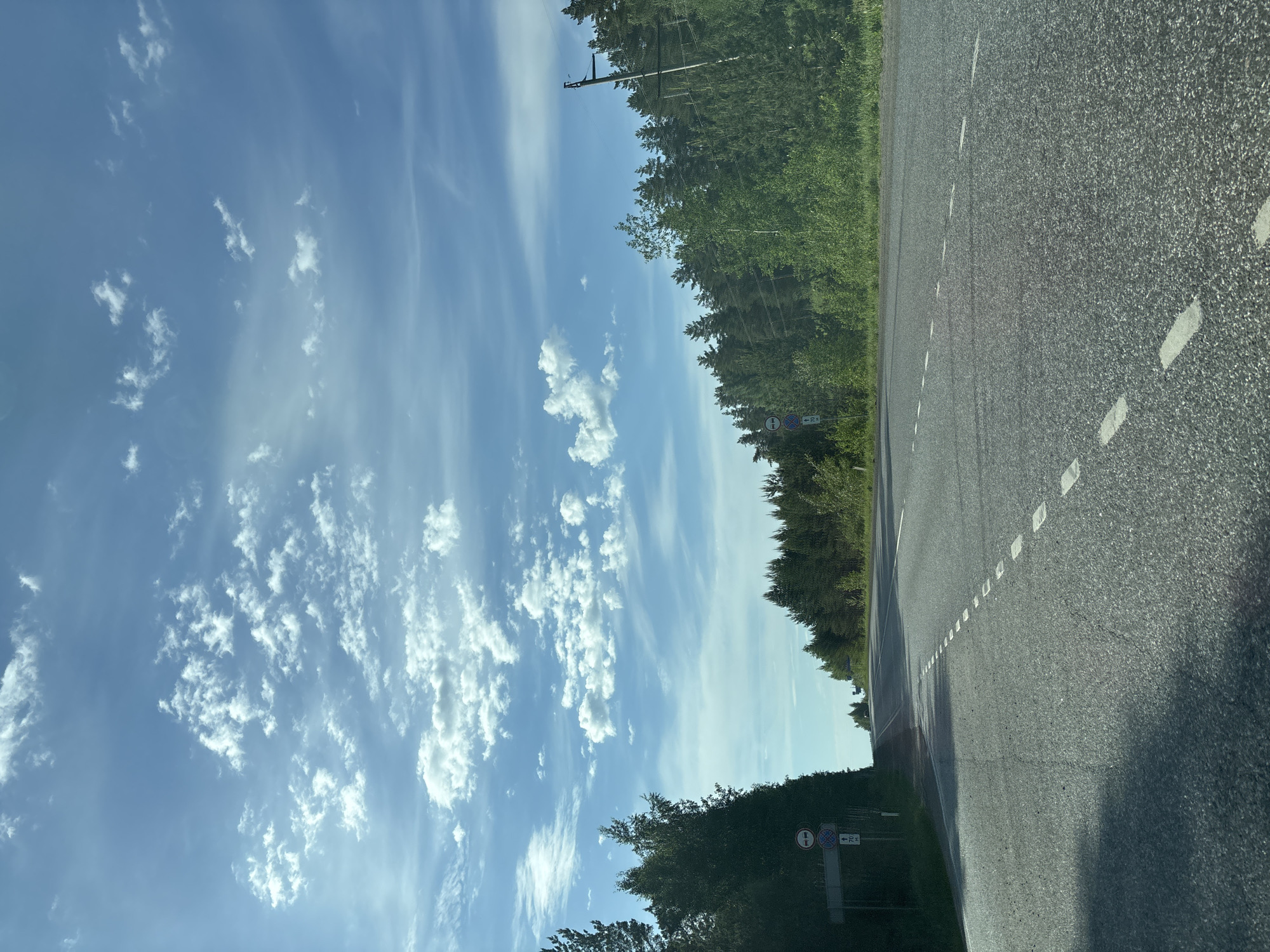 The image depicts a road in an unspecified location, with a clear blue sky filled with cirrus and cumulus clouds. The asphalt road has dotted white lines marking the lane separation. A dense green forest borders the road on one side. A road sign is visible on the side of the road, obscured by foliage. An overhead power line is seen in the background. The image is oriented sideways.