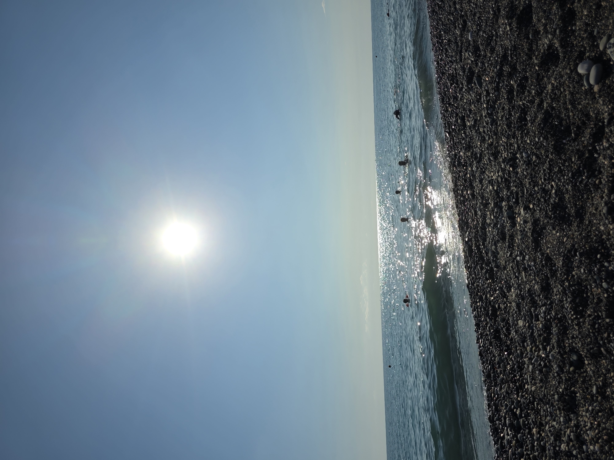 The image depicts a pebble beach on the Black Sea coast in Khostinskiy, Russia. The sun is high in the clear blue sky, creating a bright glare on the water. Several people are swimming in the sea, with only their heads visible above the surface. The beach is covered in dark, smooth pebbles. The water is calm with small waves lapping at the shore. The scene is peaceful and evokes a sense of summer leisure.