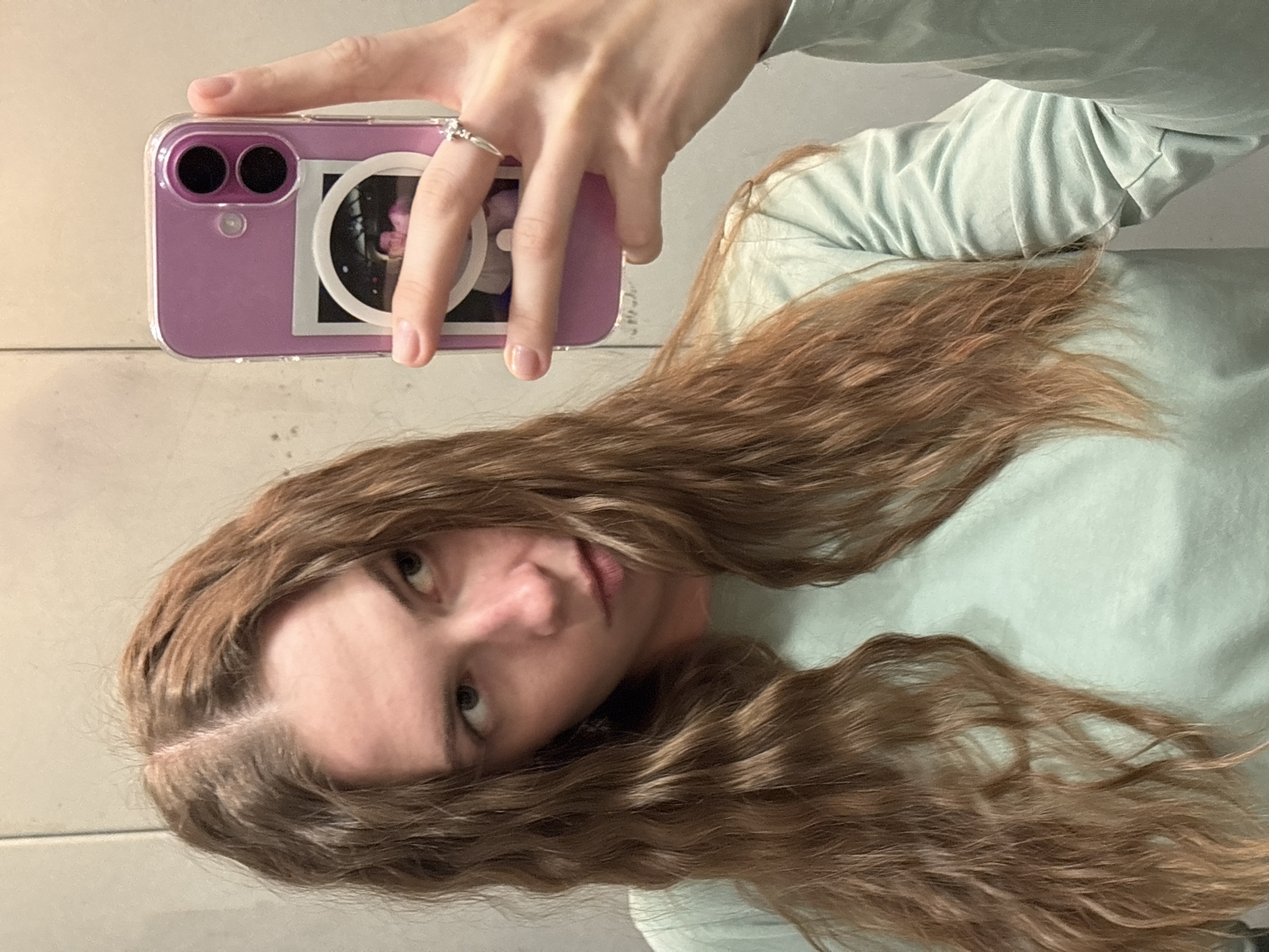A young woman with long, wavy brown hair is taking a mirror selfie indoors. She is wearing a light blue long-sleeved shirt. Her right hand, adorned with a ring, is holding a purple iPhone. The phone case has a circular sticker with a photo of a person inside. The background appears to be a plain, light-colored wall, possibly in a fitting room or a simple room. There is no indication of the city of Perm, Russia in the image.
