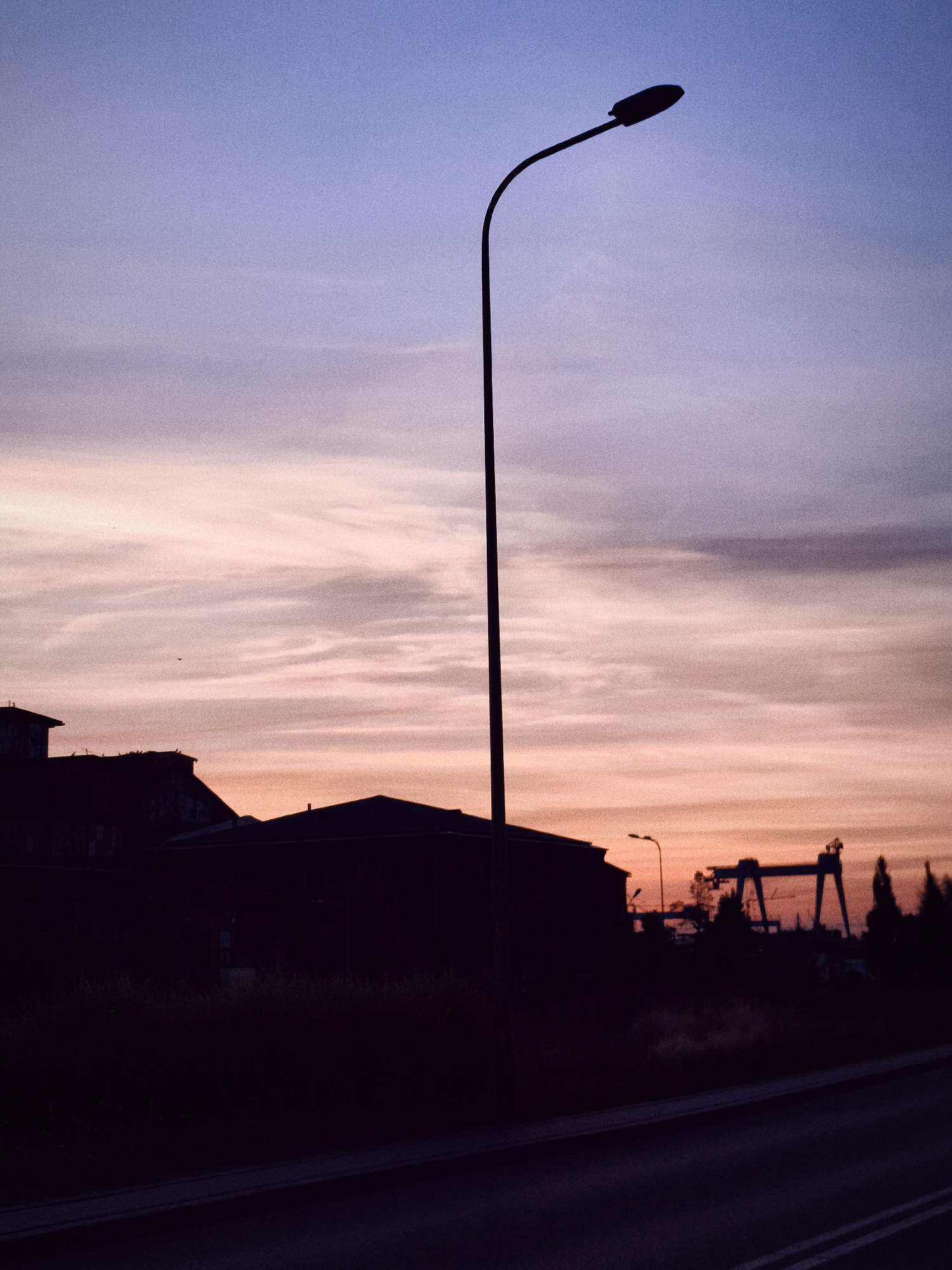 A silhouetted streetlamp stands prominently against a dusky sky in Gdańsk, Poland. The sky displays a gradient of colors, transitioning from a pale lavender at the top to soft hues of pink and orange near the horizon, indicative of twilight. In the background, the dark silhouettes of industrial buildings and structures, possibly part of the Gdańsk Shipyard, are visible. A large gantry crane stands to the right, a common sight in shipyard environments. The foreground shows a dark road with faint white lines. No people or animals are discernible in the image. The overall atmosphere is quiet and industrial, capturing the transition from day to night in a maritime city.