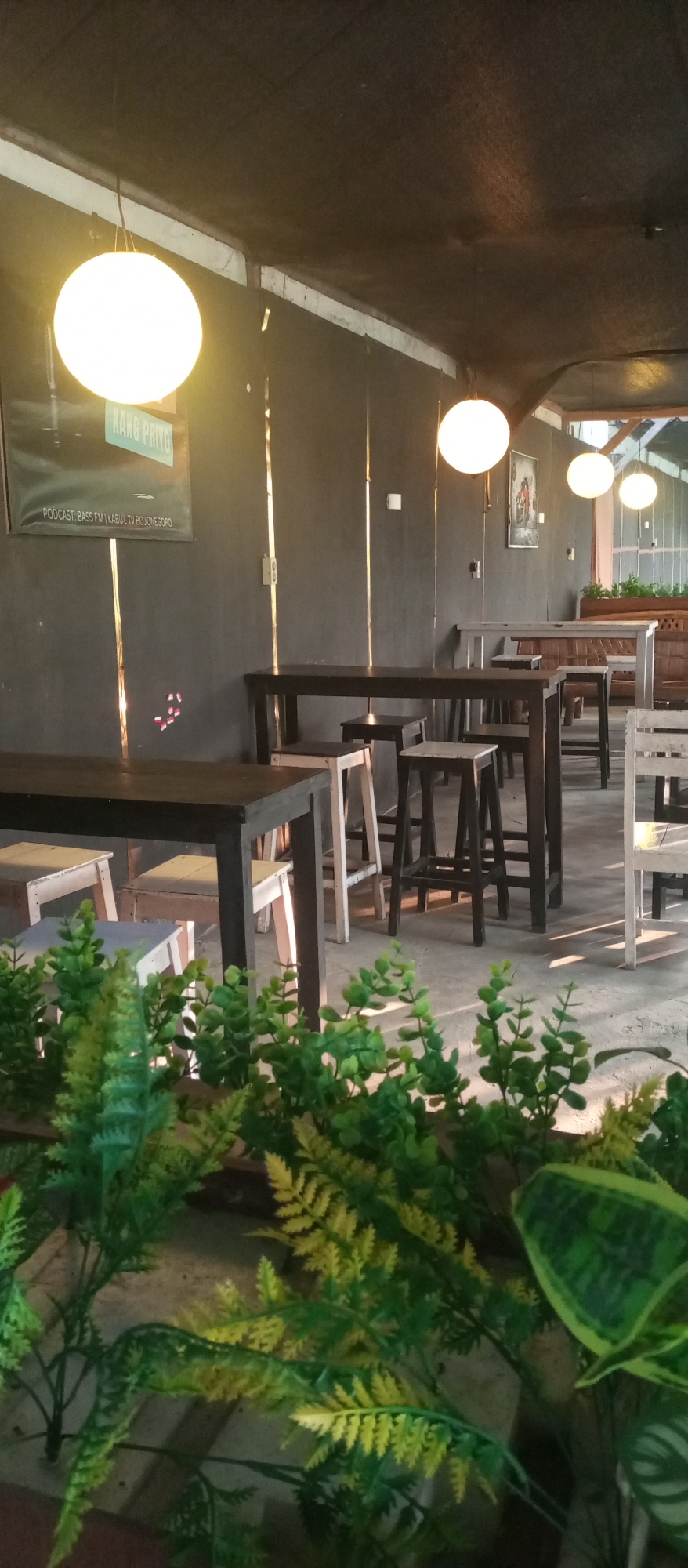 The image depicts an indoor seating area, likely a cafe or restaurant, with a rustic and dimly lit ambiance. In the foreground, lush green artificial plants with fern-like fronds are prominent, creating a naturalistic border. Beyond the plants, the scene reveals several dark wooden tables of varying heights, some paired with wooden stools. The tables are spaced to accommodate patrons, suggesting a dining or gathering space.
The walls are a dark, textured grey, accented with vertical metallic strips that reflect light. Several round, white pendant lights hang from the ceiling, casting a warm glow. One notable element on the wall is a framed poster with text that includes "KANG PRIYO" and "PODCAST: BASS FM1 KABUL TV BOJONEGORO". This poster, along with the presence of other framed artwork on the wall, suggests a cultural or community-oriented establishment.
The flooring appears to be concrete, with shadows indicating the direction of lighting. In the background, more seating areas are visible, including what appears to be a long bench or table with a wooden backrest. The overall impression is of a casual, possibly bohemian, eatery or hangout spot. The mention of "Kedewan, Indonesia" in the prompt indicates the geographical context of the image, placing this establishment within that locale. There are no people visible in the frame. The scene is static and appears to be captured during daylight hours, given the natural light filtering in from an unseen source in the distance.