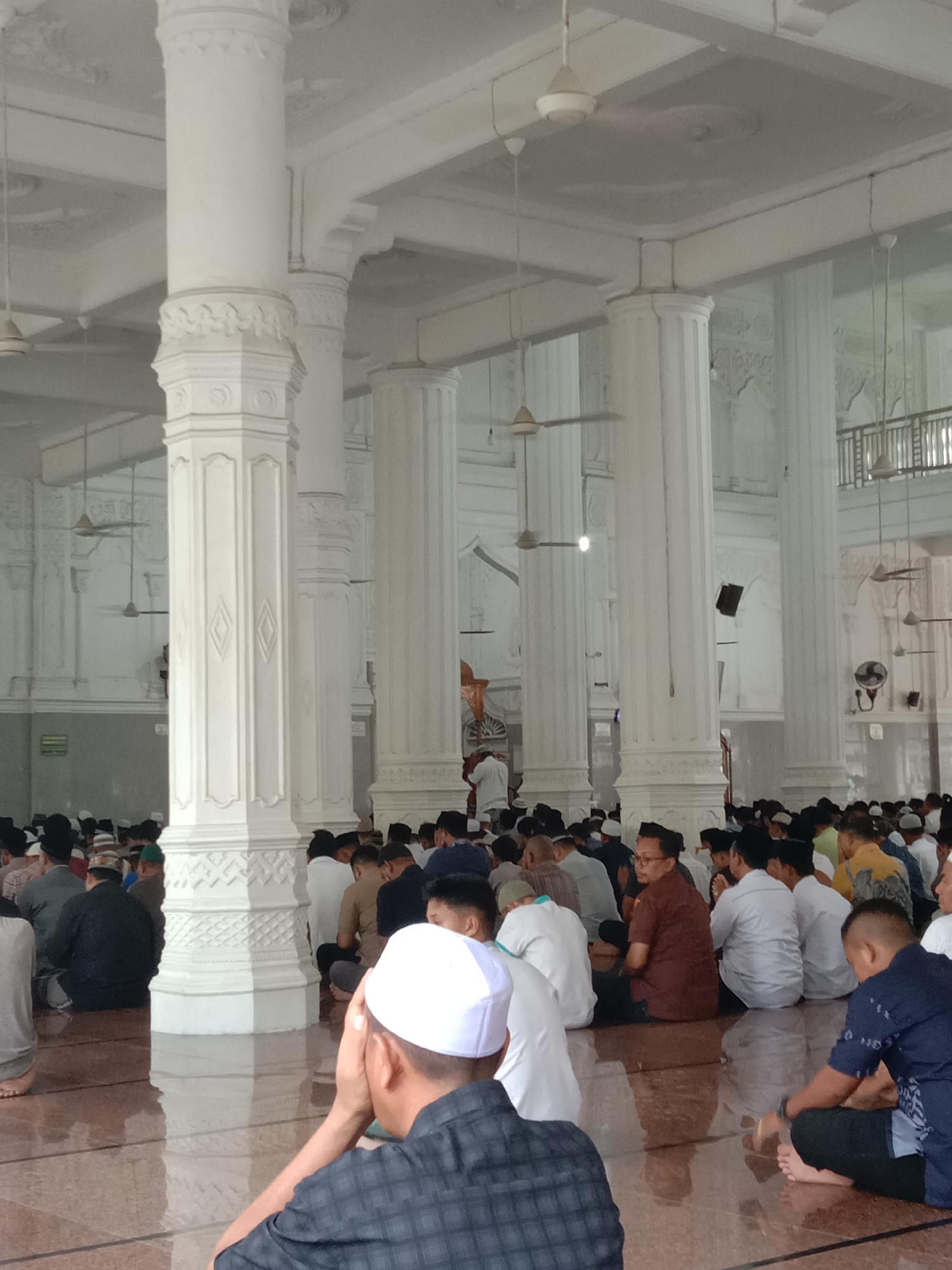 The image depicts the interior of a mosque, likely in Lhokseumawe, Indonesia, filled with a large gathering of men seated on the floor in prayer. The men are predominantly dressed in traditional Muslim attire, including various caps and long-sleeved shirts. The mosque's architecture features large, ornate white pillars and a high, white ceiling with multiple ceiling fans. The floor appears to be made of polished wood or tile, reflecting the ambient light. In the foreground, several men are visible from behind, engaged in prayer, with one man in a white cap covering his face with his hands. The scene conveys a sense of communal worship and religious devotion.