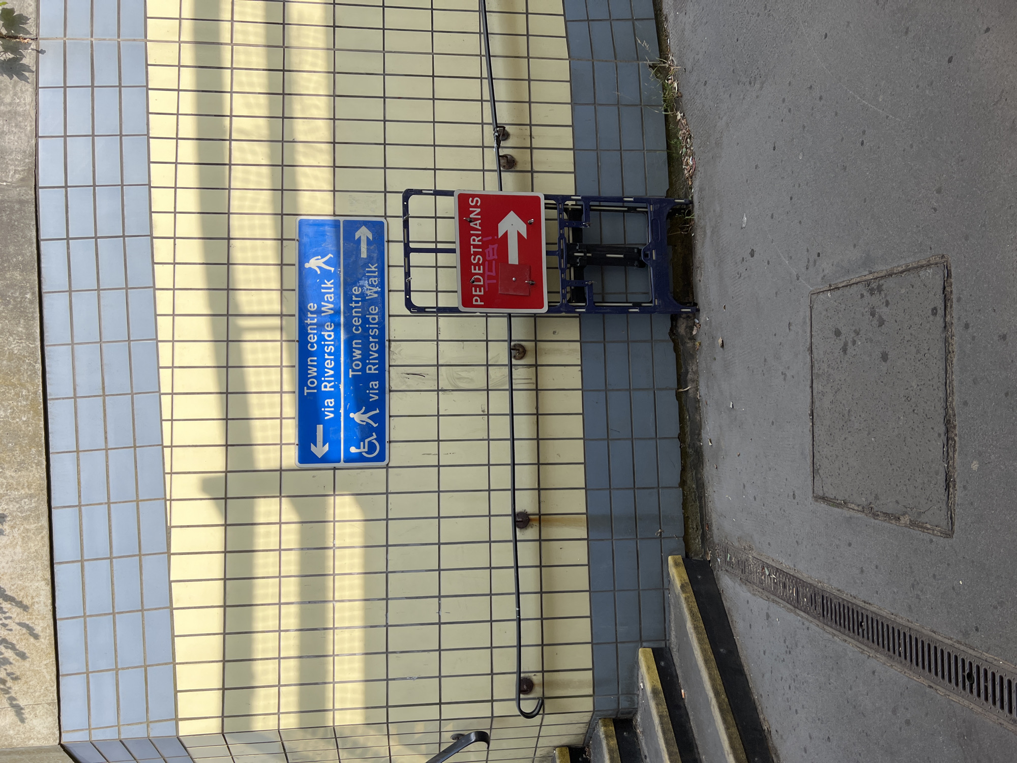 The image shows a section of a tiled wall with two blue directional signs and a red temporary sign on a blue stand. The wall is tiled with pale yellow tiles in the lower section and pale blue tiles in the upper section.
The top blue sign has a left-pointing arrow and text that reads "Town centre via Riverside Walk" with a pictogram of a person walking.
The second blue sign has a right-pointing arrow and text that reads "Town centre via Riverside Walk" with a pictogram of a person in a wheelchair.
In front of the signs is a blue metal folding A-frame stand holding a red sign. The red sign has white text that reads "PEDESTRIANS" above a white arrow pointing to the right. There is some red graffiti or writing above the arrow on the red sign.
To the left of the wall are concrete steps with black and yellow edging. A metal handrail runs along the wall above the steps. The ground in front of the wall is paved concrete with a metal drain grate and a square concrete utility cover. Some sparse vegetation is growing at the base of the wall.
This scene is located in Guildford, United Kingdom, specifically at the entrance to the underpass beneath the railway line near Guildford Station, leading to the High Street and Riverside Walk.