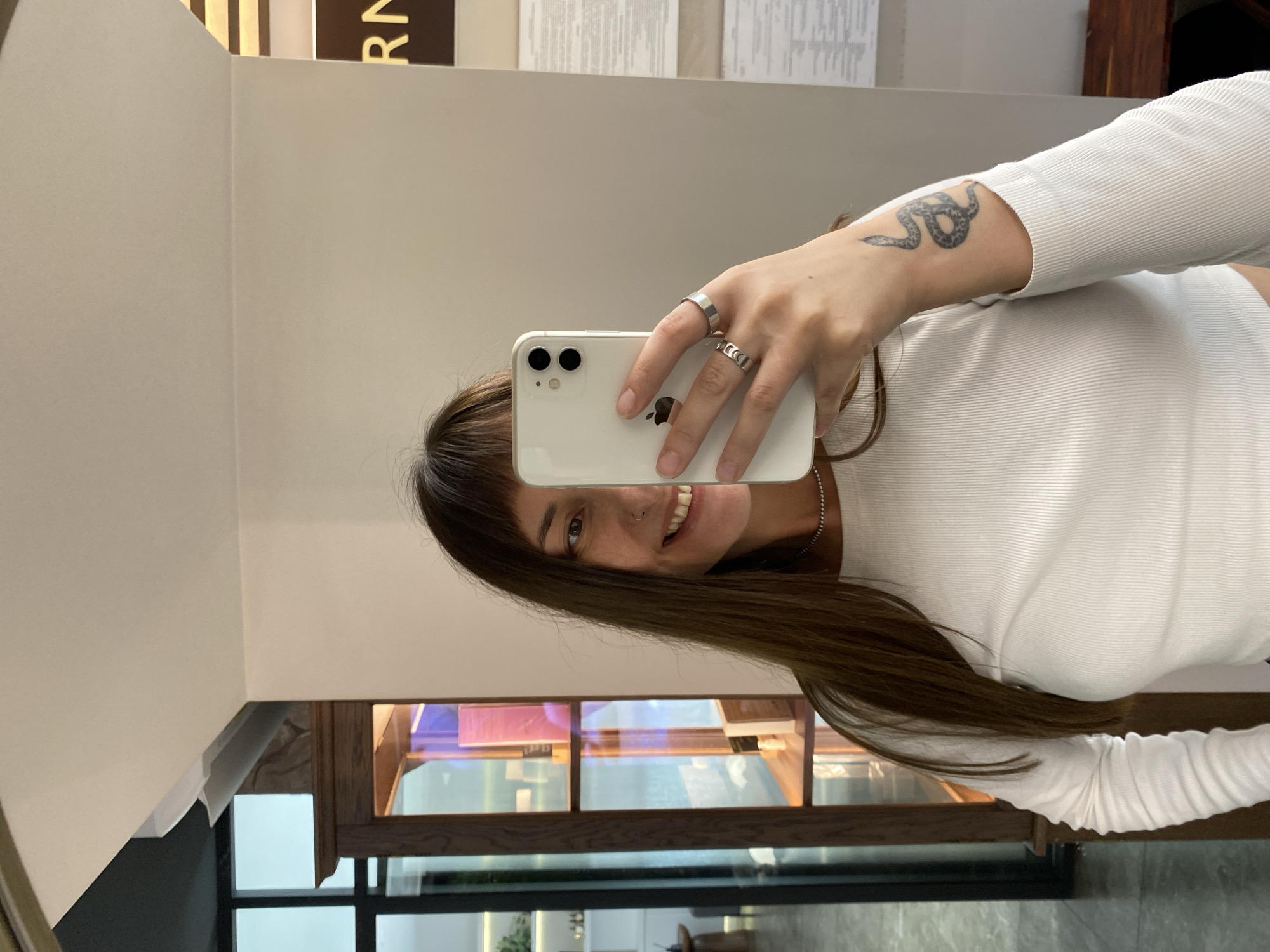 A woman with long brown hair and bangs smiles at the camera while holding a white smartphone in her right hand, taking a selfie in a mirror. She is wearing a white, long-sleeved, ribbed shirt and several silver rings on her fingers. A snake tattoo is visible on her left forearm. The background appears to be an indoor space with a wooden cabinet filled with books to the left, and a cream-colored wall to the right. The image does not contain any information that would allow for identification of the location as Votkinsk, Russia.