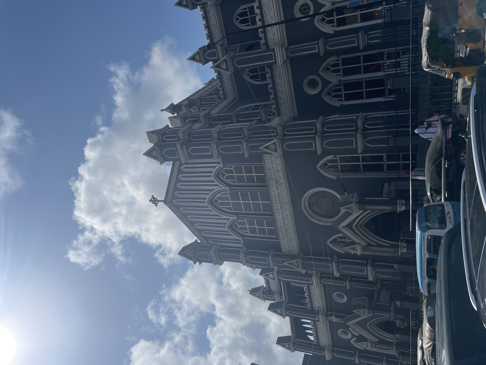 The image shows a partial view of the Cathedral Church of Christ in Lagos, Nigeria. The cathedral is a large grey building with white trim and arched windows. The architectural style appears to be Gothic Revival. The sky is visible in the background, with white clouds and blue patches. Part of the street with cars and auto rickshaws are visible on the right side.