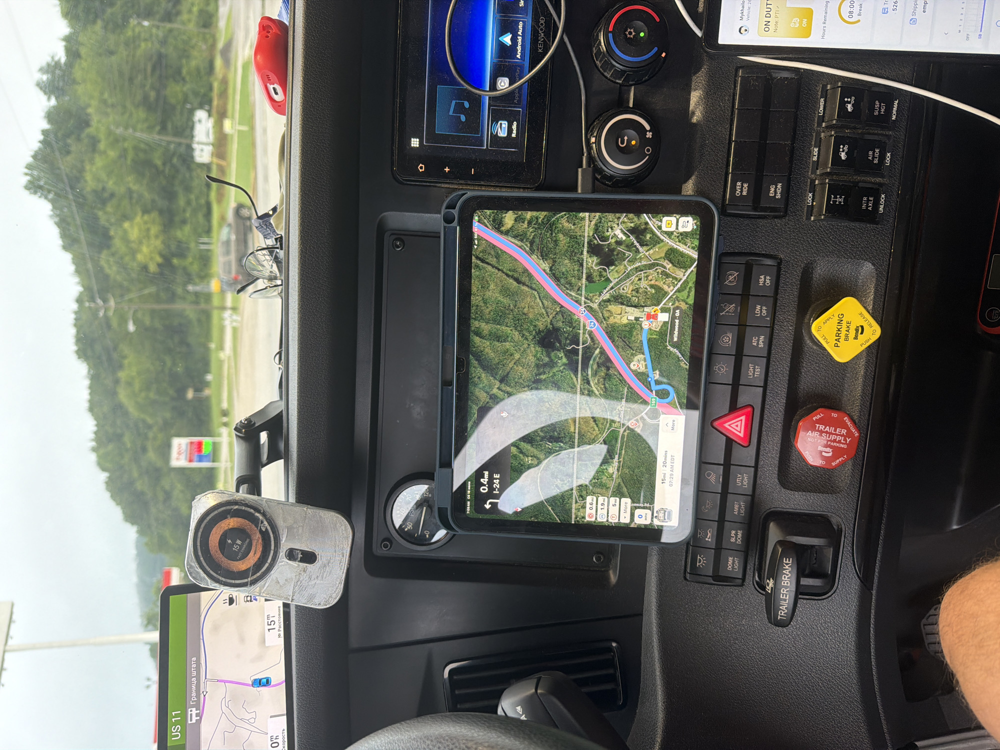 The image shows the interior of a truck's cabin, likely a semi-trailer truck, focusing on the dashboard area. A tablet mounted on the dashboard displays a GPS navigation screen with a route highlighted, showing the location as "Wildwood, GA." Various buttons and controls for the truck's functions are visible, including "TRAILER BRAKE" and "PARKING BRAKE." A secondary display, possibly a car stereo or infotainment system, is also present. In the background, through the windshield, a gas station with visible signage and trees can be seen, suggesting the truck is parked at a service station. The lighting indicates it is daytime, and the sky appears overcast. No people are clearly visible in the immediate cabin area.