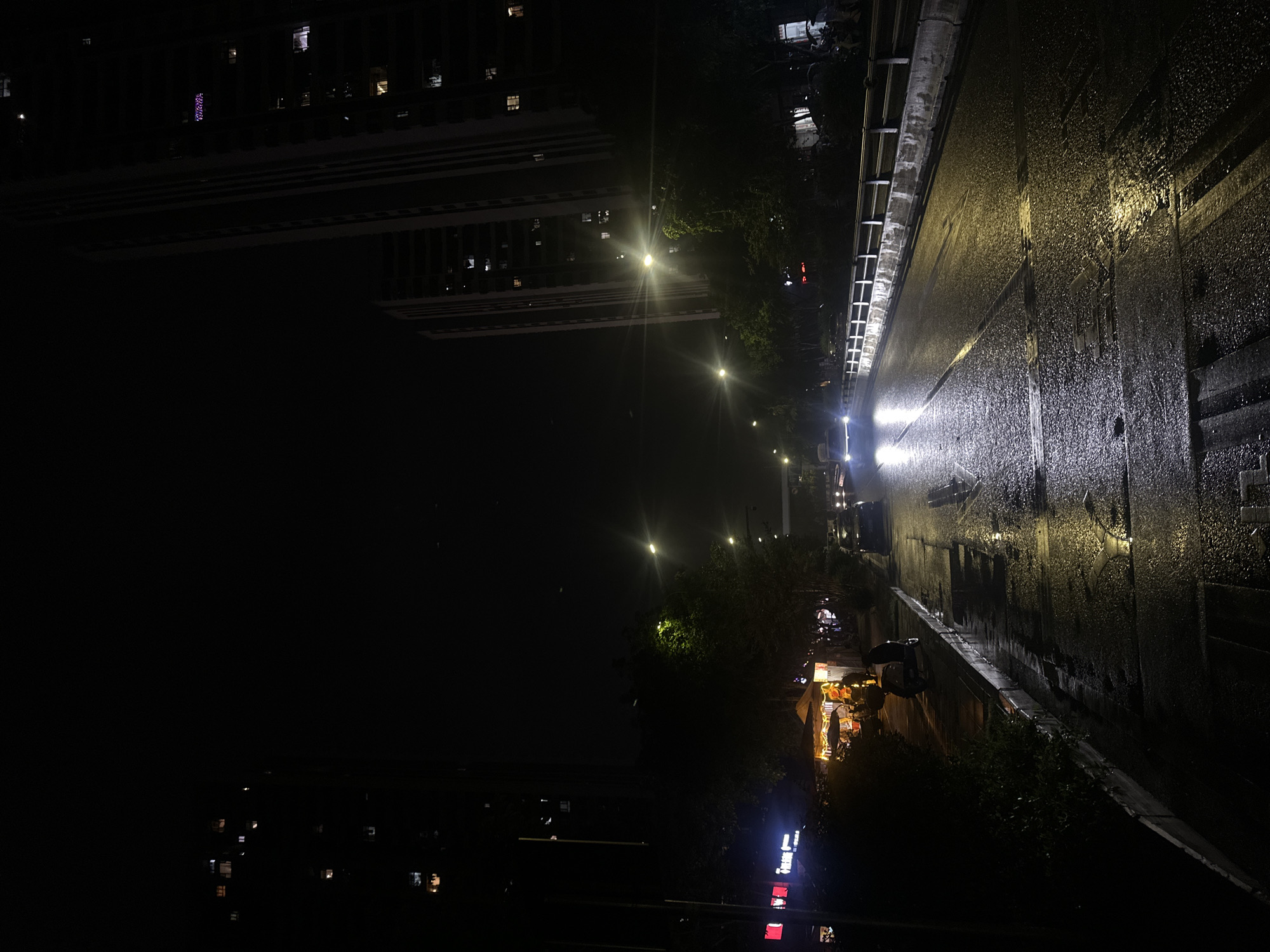 The image depicts a night scene in Chongqing High-tech Zone, China, taken from an elevated position. A wet, reflective road surface occupies the right side of the frame. Streetlights illuminate the road and the surrounding trees and buildings. On the left, tall buildings are visible, with lit windows indicating occupancy. Further down, there are shops with orange lights. At the lower end, the lights of buildings and signs indicate an urban area.