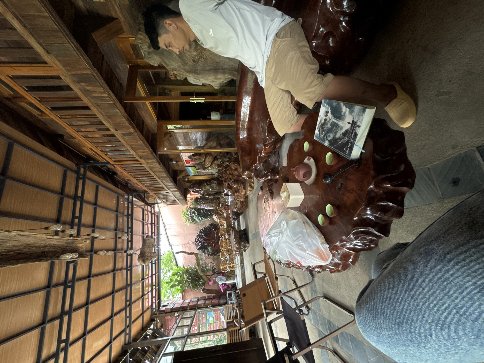 The scene depicts a man resting on a large, ornate wooden table in what appears to be an indoor/outdoor space. The man is wearing a white shirt and tan shorts, and is positioned with his head resting on a section of the table that resembles a large, polished tree trunk. On the table, next to his feet, is a book. There are also cups, a teapot, and a white plastic bag. The space is enclosed by a structure with a partially exposed roof, showcasing views of trees and greenery in the background. The location is in Bản Đêu, Vietnam.