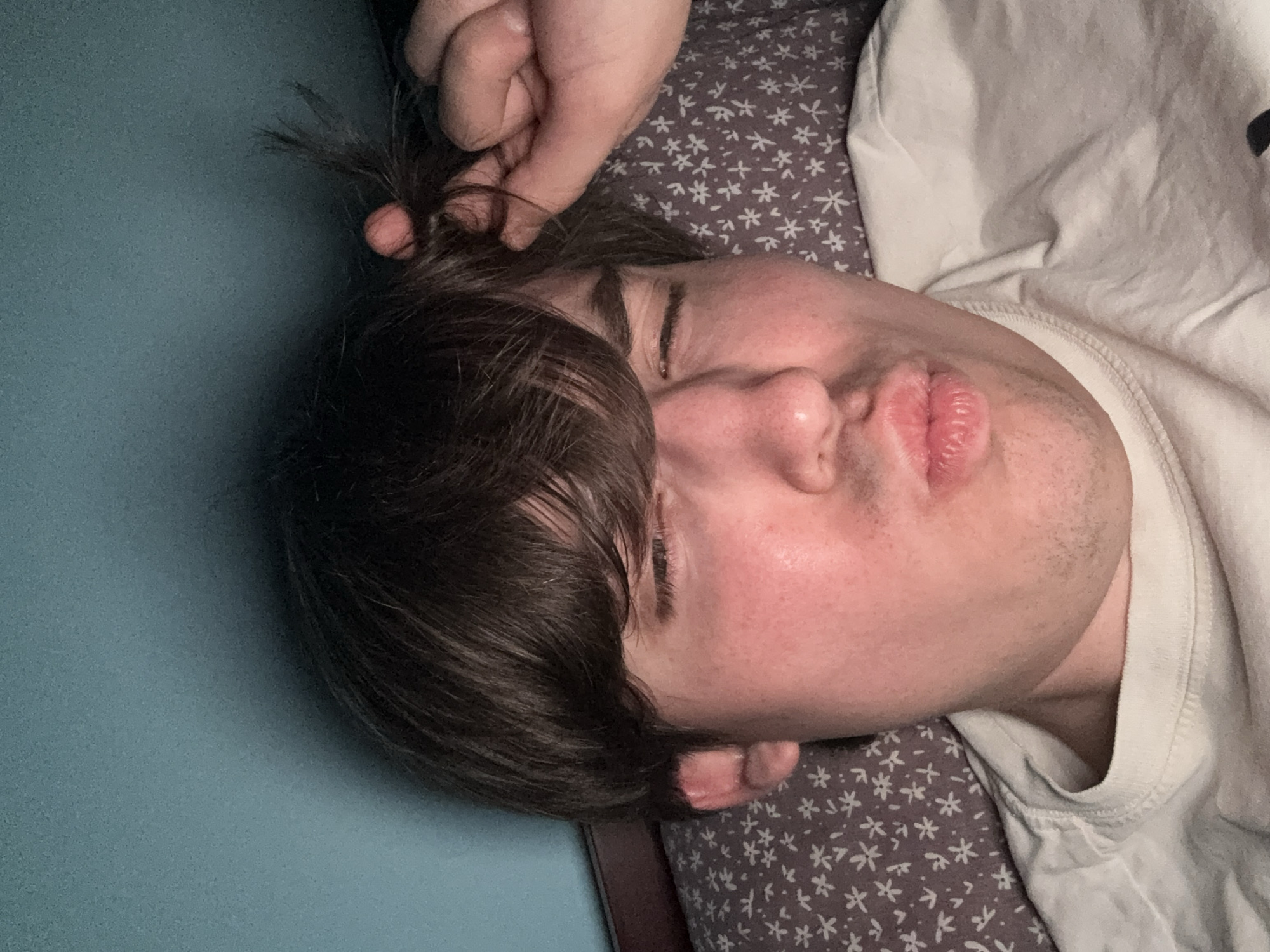 A close-up, low-angle, selfie-style photograph shows a young person with brown hair and fair skin lying down. The person is wearing a white t-shirt and their eyes are closed, with their lips puckered in a kissing expression. Their right hand is raised to their hair, holding a few strands. The person is lying on a pillow with a small white floral pattern on a grey background. The background appears to be a wall painted a teal or blue-green color. The lighting suggests artificial light, possibly from a phone or overhead lamp, creating some shadows and highlights on the person's face and the pillow. The image does not provide any information that would allow for the determination of its location in the city of Dnipro, Ukraine.
