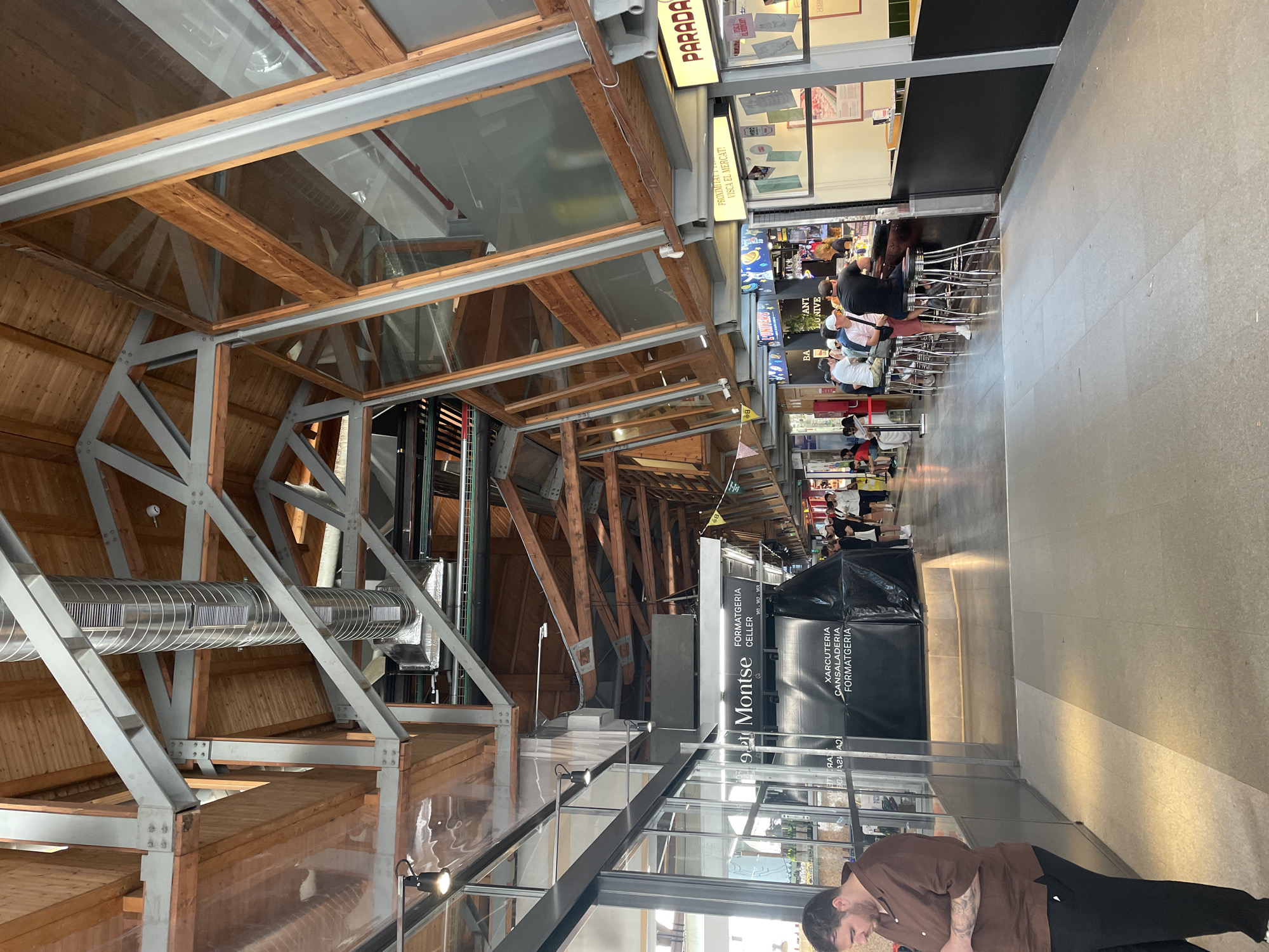 The image depicts the interior of a market hall with a high, exposed wooden and metal beam ceiling. Large windows line the upper walls, allowing natural light to enter. Below the ceiling, ventilation ducts are visible. The market appears to be bustling with activity.
In the foreground to the left, a man wearing a brown shirt and black pants stands facing away from the camera, looking at his phone.
Further into the hall, various market stalls are set up. One prominent stall has black signage with white text that reads "Montse FORMATGERIA CELLER" and below that, "XARCUTERIA CANSALADERIA FORMATGERIA". A large black tarp covers part of this stall.
To the right, a bar area is set up with several patrons seated on stools. People are standing and interacting at other stalls. The floor is tiled, with a distinct line of light and shadow marking a path.
In the far right of the image, a stall with a yellow sign that says "PARADA" is visible.
The overall impression is of a lively indoor market, likely in Barcelona, Spain, given the signage which includes Catalan phrases like "VISCA EL MERCAT" (Long live the market).