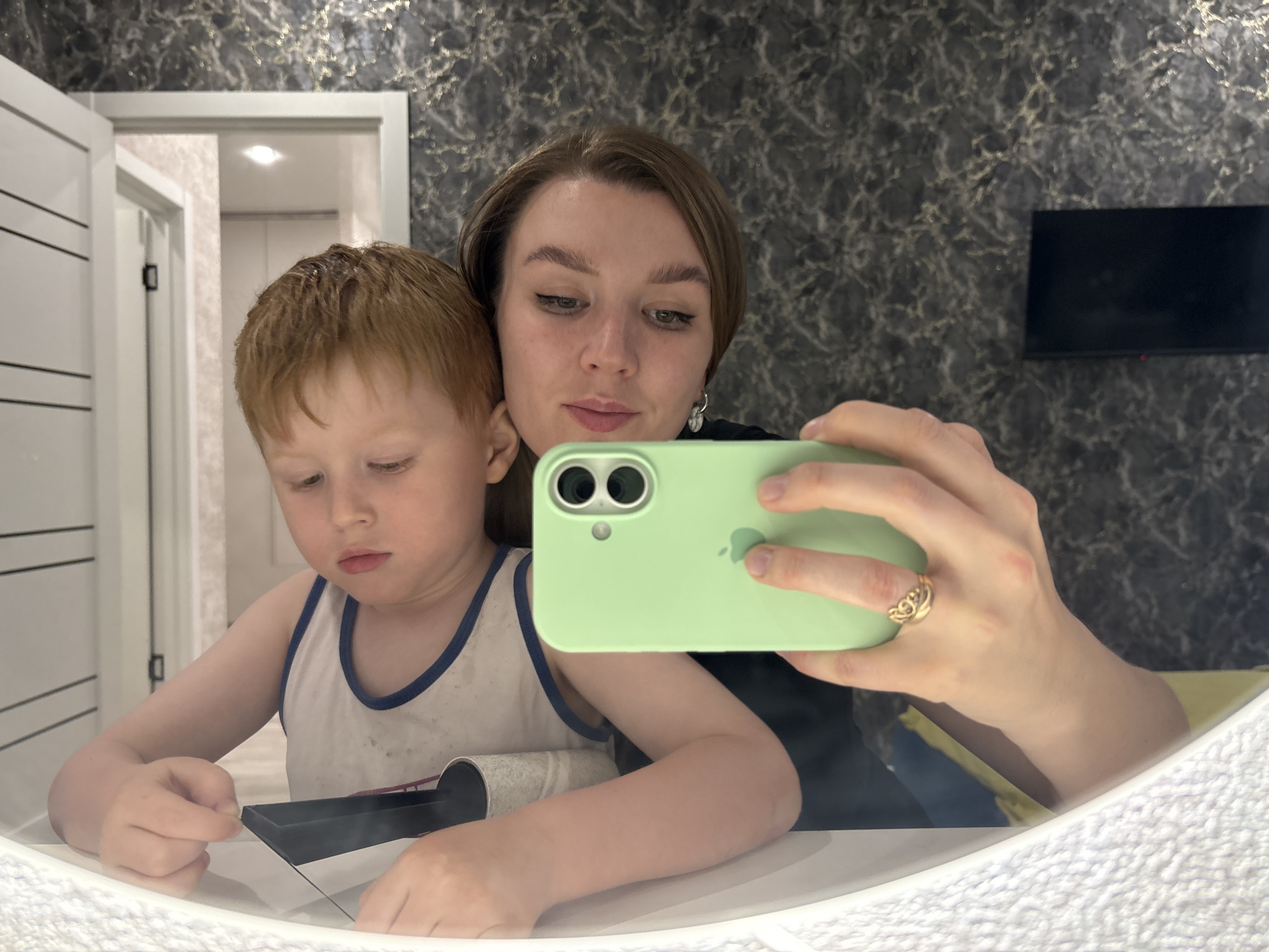 A young woman and a young boy are visible in a mirror selfie taken indoors. The woman holds a light green smartphone with two camera lenses in her right hand. She has short brown hair and wears eyeliner. She also wears a gold ring on her right hand and earrings. The boy, who is fair-skinned with red hair, wears a white tank top with blue trim. He is looking down at a black object on the counter in front of him. A white lint roller is also visible on the counter. The background includes a wall with a dark, marble-like pattern, a flat-screen television, and a partially open white door leading into another room. The photograph was taken in Izhevsk, Russia.