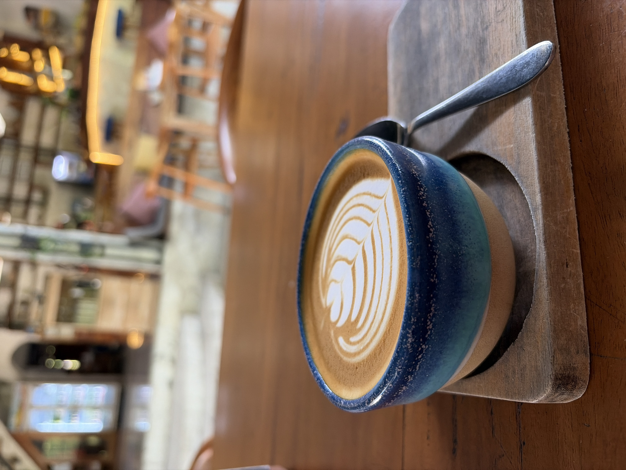 A close-up, eye-level shot shows a cup of latte art coffee on a wooden table at a cafe in Kubu, Indonesia. The coffee is in a blue and tan ceramic bowl with a spoon resting on a small wooden board. The background is blurred, showing tables, chairs, and lights, suggesting a bustling cafe environment. The lighting is soft and natural, creating a warm and inviting atmosphere.