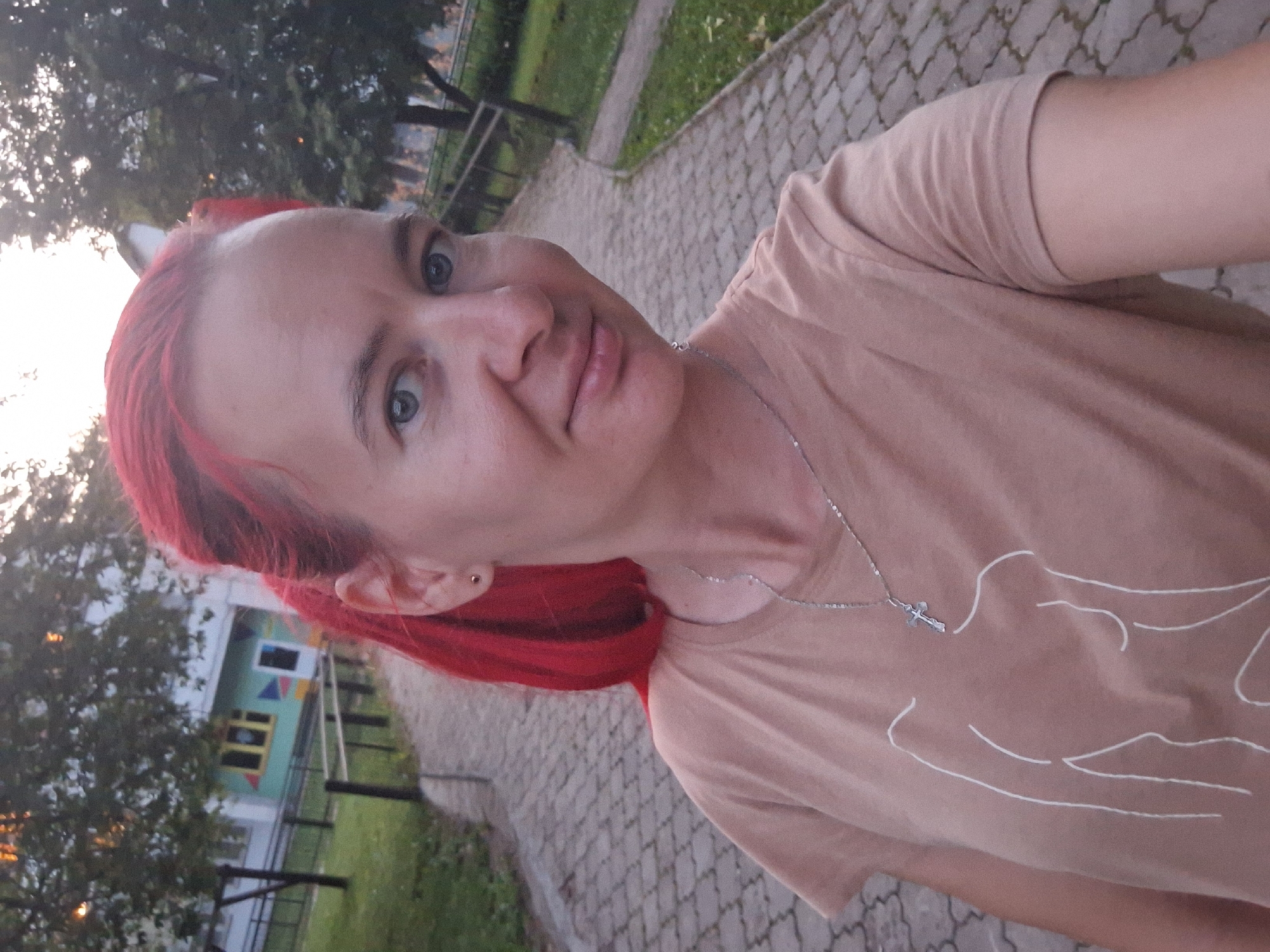 A woman with bright red hair, wearing a light brown t-shirt with a white line drawing of a torso and a silver necklace with a cross pendant, stands on a cobblestone path in an outdoor setting. She is looking at the camera and smiling slightly. Behind her, a building with colorful geometric decorations is visible, along with trees and a railing. The scene appears to be in a park or residential area, likely in the city of Atamanovka, Russia, given the context. The lighting suggests it is late afternoon or early evening.