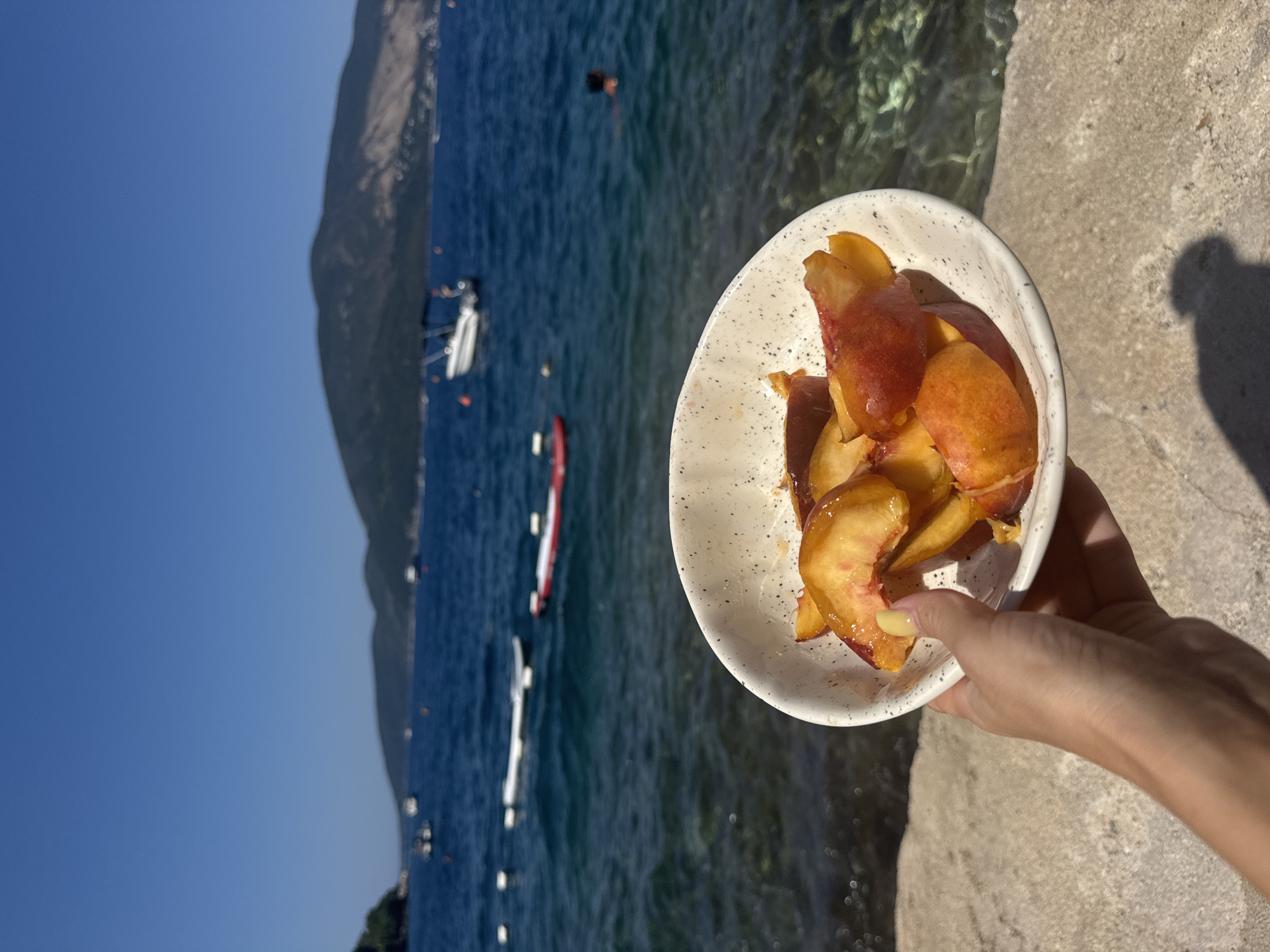 A hand holds a white bowl of sliced peaches in the foreground, with a body of blue water and a distant, tree-covered mountain range in the background. Several boats and floating devices are visible on the water, and a person is swimming. The scene appears to be a scenic overlook on a sunny day, likely near a beach or coastline. The location is not specified beyond "Unknown, Unknown."