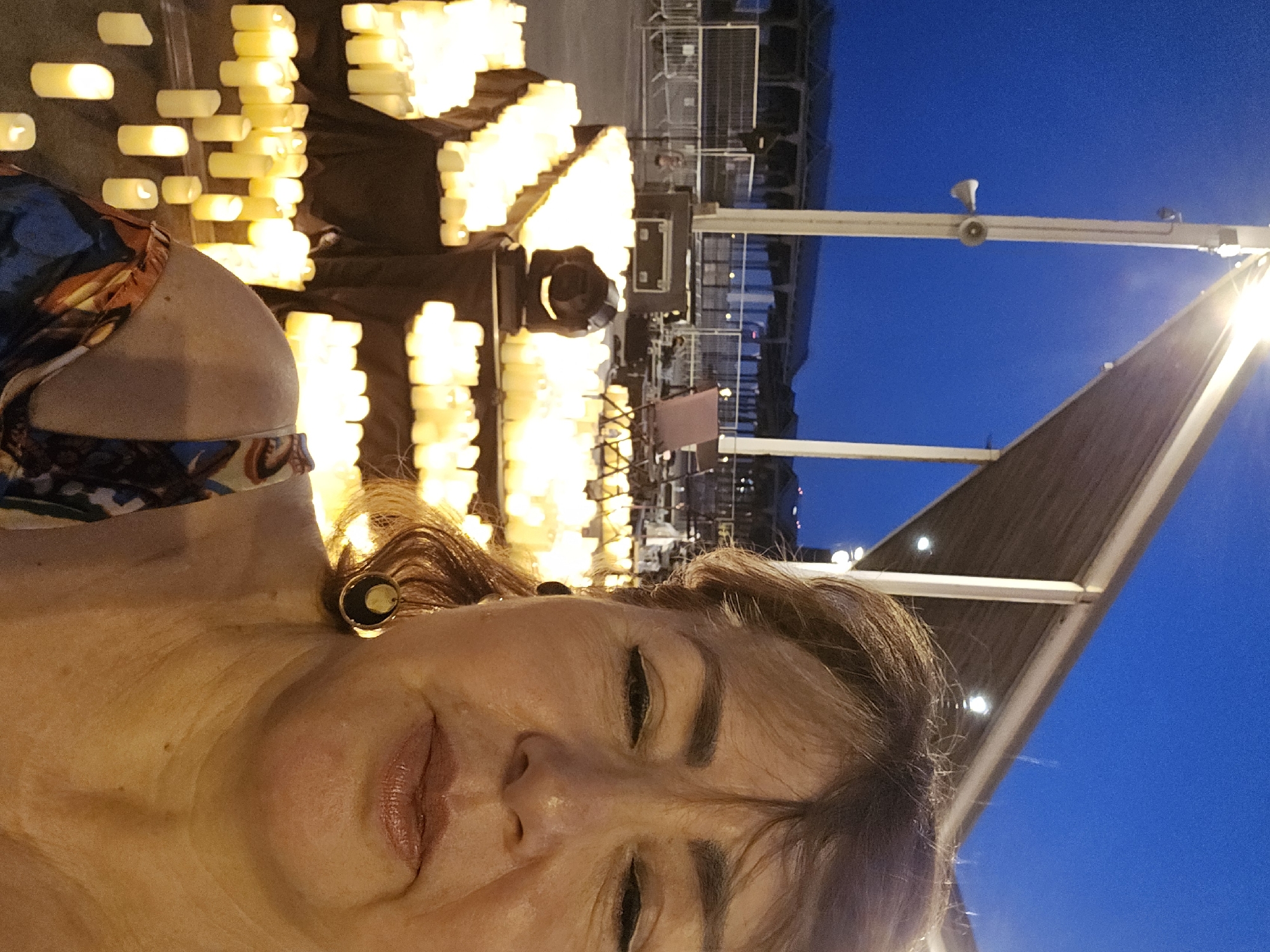 A medium shot selfie shows a woman in the foreground, looking towards the camera. Her eyes are closed and her lips are slightly parted. She is wearing a patterned dress and large, circular earrings. Behind her, a large display of lit candles is set up on tables and on the ground. The background features a clear blue sky at dusk, a lighting rig, and what appears to be a stage with equipment. The location is the Parc del Litoral in Sant Adrià de Besòs, Spain, where a vigil of remembrance was held.