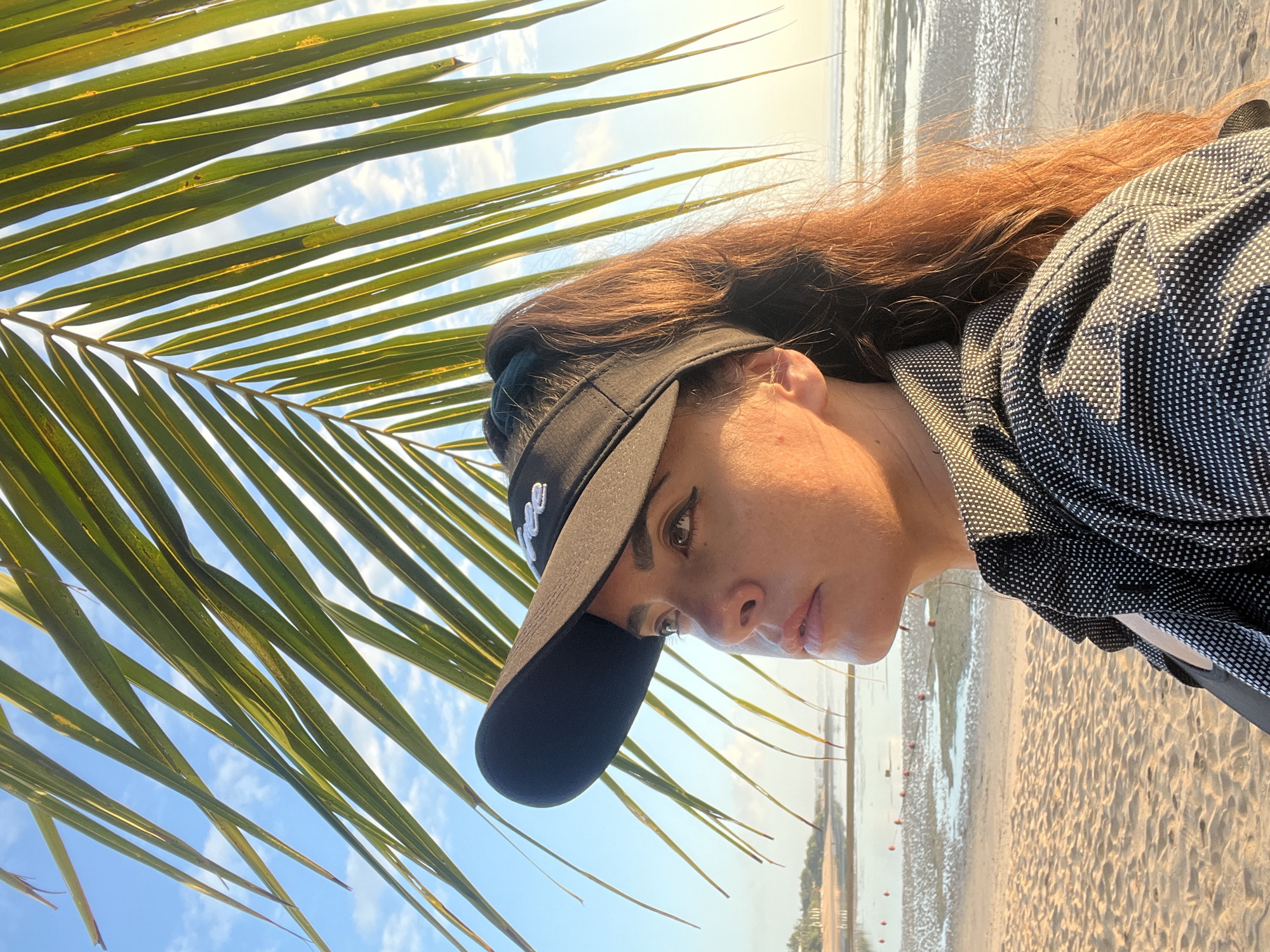 The image depicts a medium shot of a woman standing on a sandy beach, likely in Kuta Selatan, Indonesia, with a palm frond in the foreground. The woman is wearing a black visor with a white logo and a black and white patterned shirt over a black garment. Her hair is dark brown and tied back. She is looking to her left, with her gaze directed slightly upwards. The background shows a calm body of water, a wet sandy shore with small buoys, and a clear sky with scattered clouds, illuminated by what appears to be morning or evening sunlight. The overall scene suggests a relaxed atmosphere by the sea.