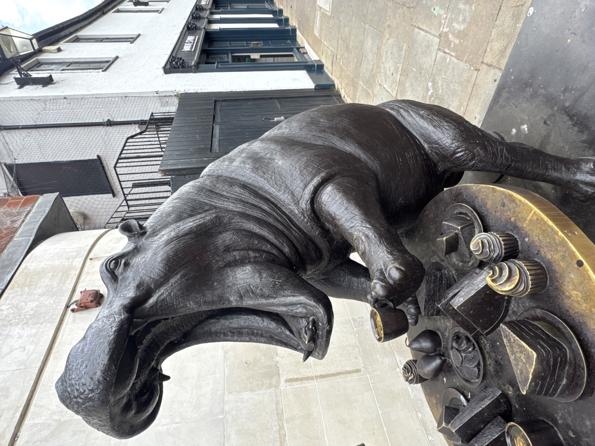A bronze statue of a hippopotamus is depicted in a public space, possibly a street or pedestrian area. The hippo is positioned on its hind legs, leaning over a table laden with bronze sculptures of food items such as cakes, fruit, and what appear to be crackers or biscuits. The hippo's mouth is open wide, as if about to eat, and it is holding a teacup in one of its front paws. The statue is dark brown in color with some lighter, possibly golden, highlights, particularly on the table and some of the food items.
In the background, a white building with dark-trimmed windows and doorways is visible, with a sign that reads "WHITE STUFF". There is a metal railing and a wooden gate to the left of the building. A paved walkway made of stone slabs runs alongside the building. The overall scene suggests an outdoor art installation in an urban environment. This installation is located on Bury Street in St. James's, London, United Kingdom, in front of the White Stuff clothing store.