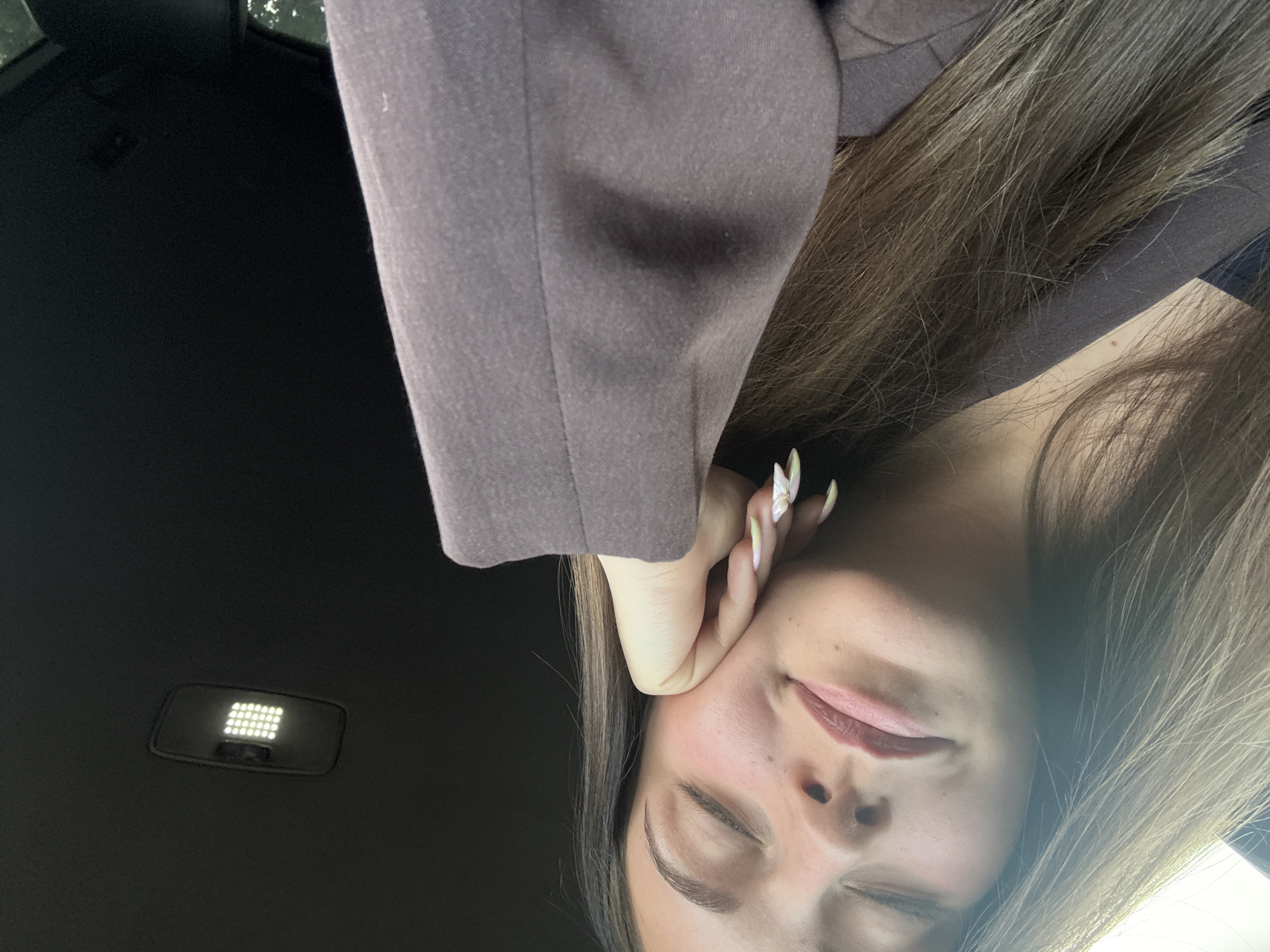 A close-up, tilted image shows a person in a car. The person, possibly female, is wearing a brown blazer. Her right hand is supporting her face near the ear. She has long, light brown hair and painted nails. The car's interior is visible in the background. An overhead light is visible. It is not possible to identify the specific location in Burgas, Bulgaria from the provided image.