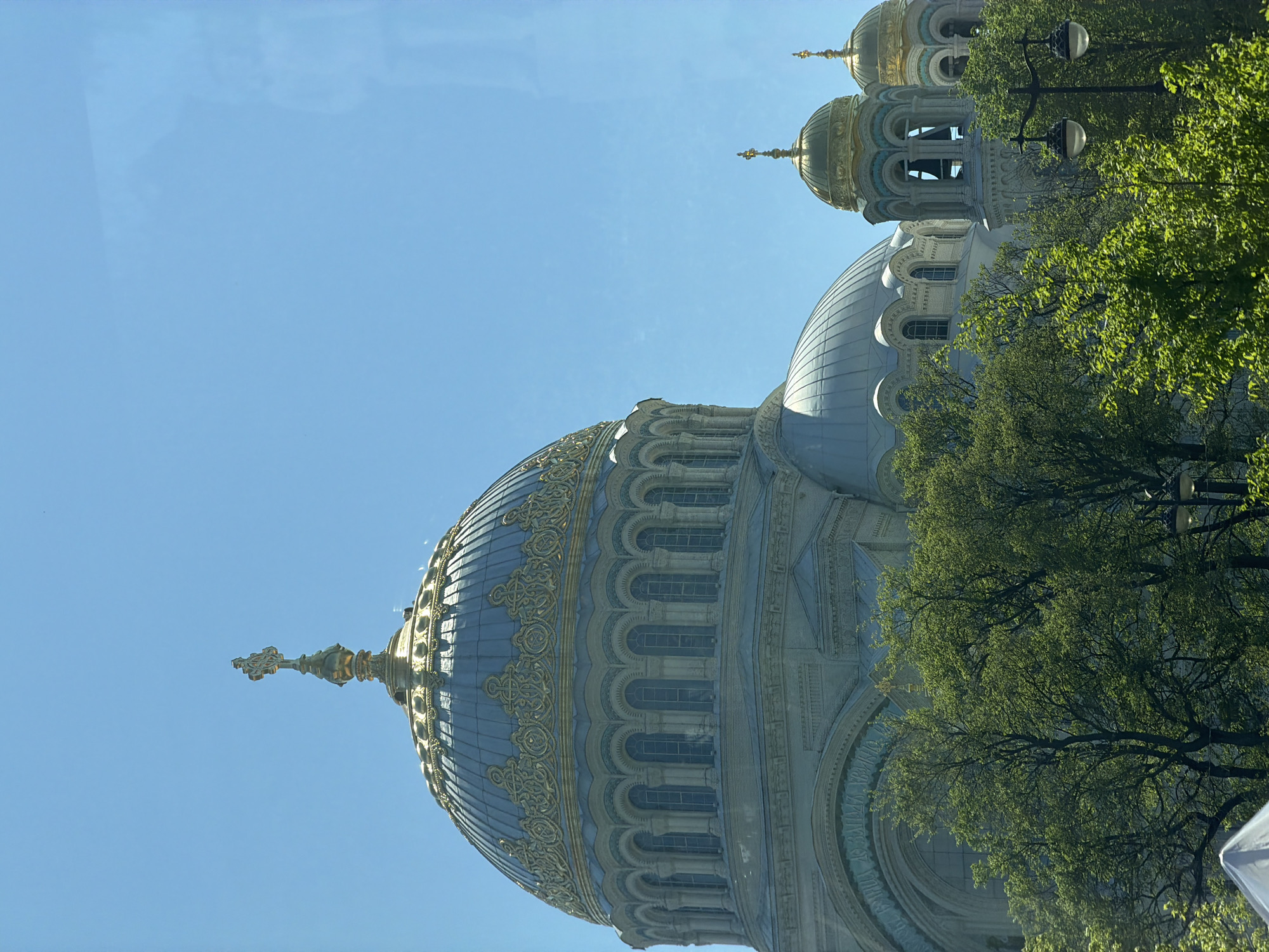 The image shows a view of the Naval Cathedral of Saint Nicholas in Kronstadt, Russia. The dominant feature is the large, blue-grey dome of the cathedral, elaborately decorated with gold detailing. To the right and slightly behind the main dome, a smaller, similarly styled dome is visible. Green trees partly obscure the lower part of the cathedral, with the branches and leaves appearing in the foreground. A couple of street lights can be seen amidst the trees. The sky is clear and blue. The image is taken from a low angle.